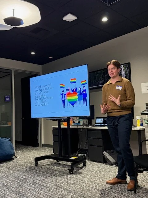 Non-binary person giving a presentation with a large screen displaying a slide about LGBTQ+ support and advocacy at a professional event.