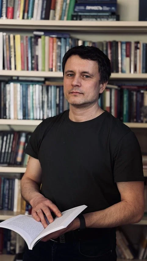 Man standing in front of a bookshelf, holding an open book.