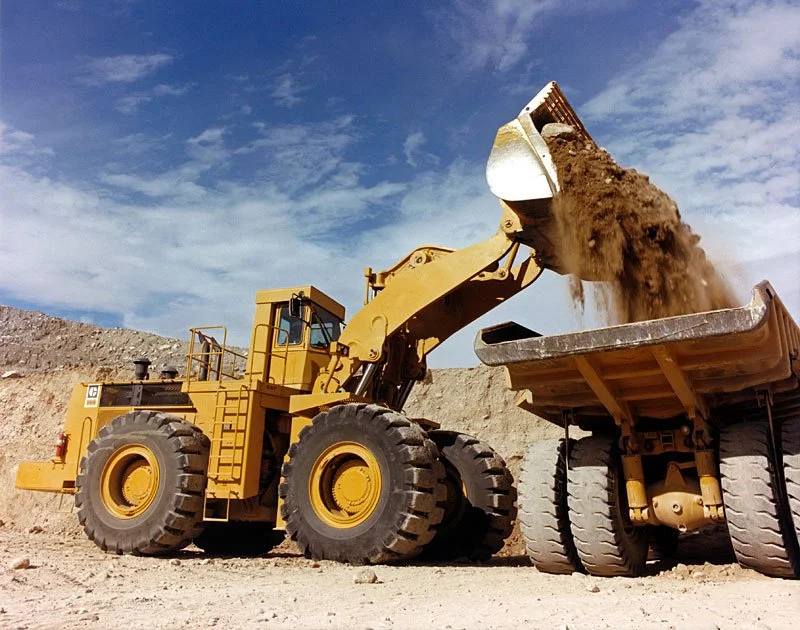 Yellow front loader dumping dirt into a large dump truck at a construction site under a cloudy sky.