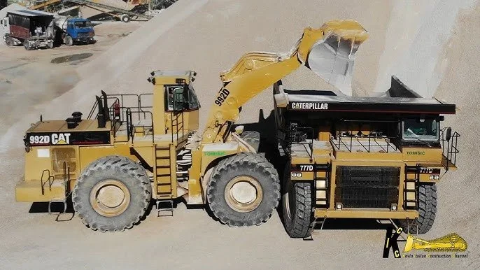 A large yellow Caterpillar 992D front loader with a scoop, and a black and yellow Caterpillar 777D dump truck in a construction site with dirt piles.