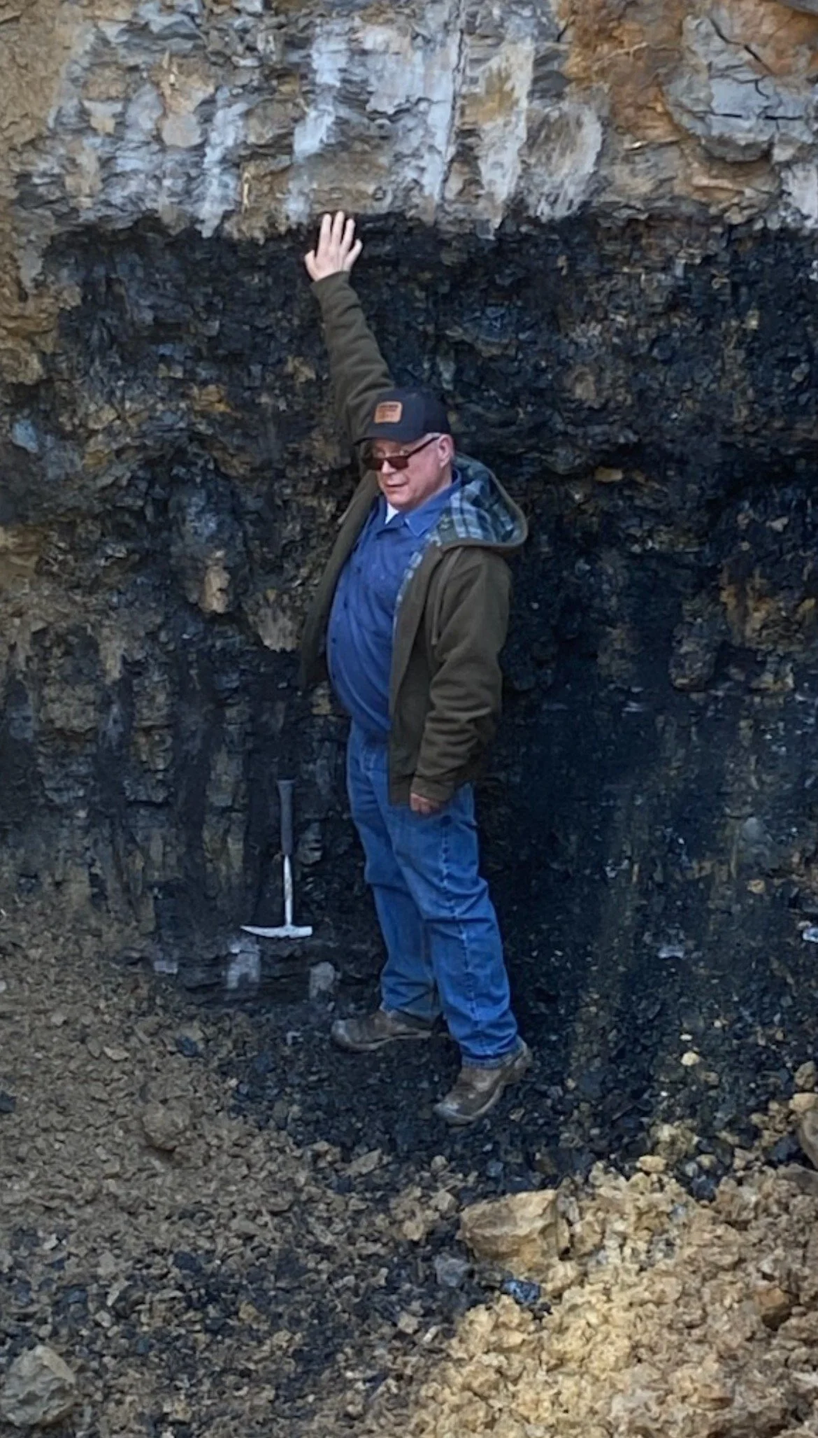 A man wearing sunglasses, a baseball cap, a blue shirt, a jacket, and jeans stands next to a rock formation with dark layers of volcanic rock. He is touching the rock with his left hand and holding a geological hammer with his right hand.