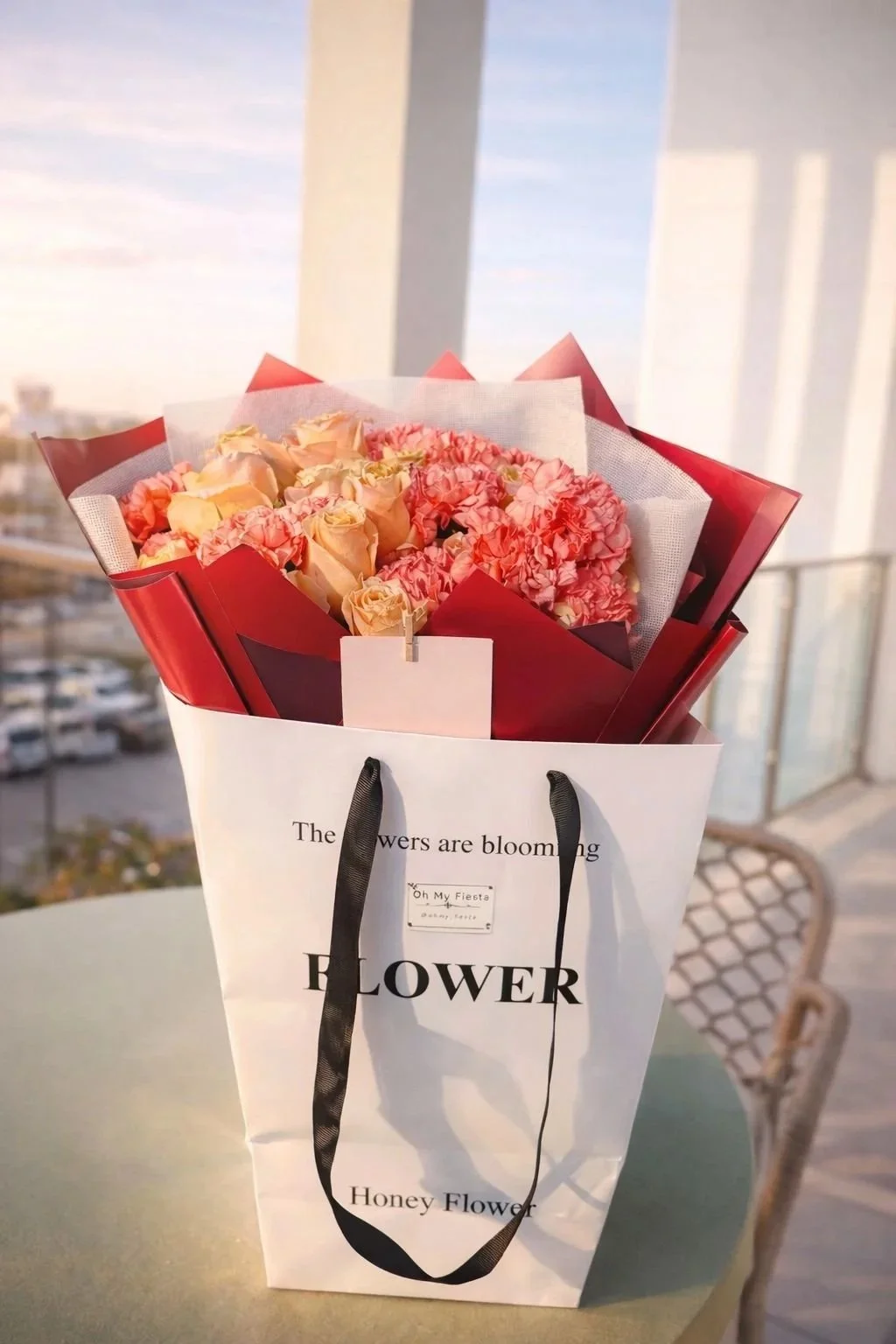 A bouquet of pink and cream roses and hydrangeas in paper wrapping, inside a white gift bag with black handles, on a table outside near a railing, with a cityscape in the background during sunset.