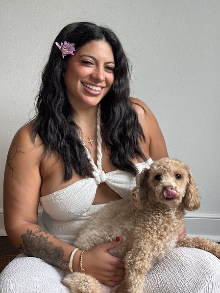 Smiling woman with dark wavy hair, wearing a white top with a knot and a pink flower in her hair, holding a tan curly-haired dog with its tongue out, sitting on the floor against a plain gray wall.
