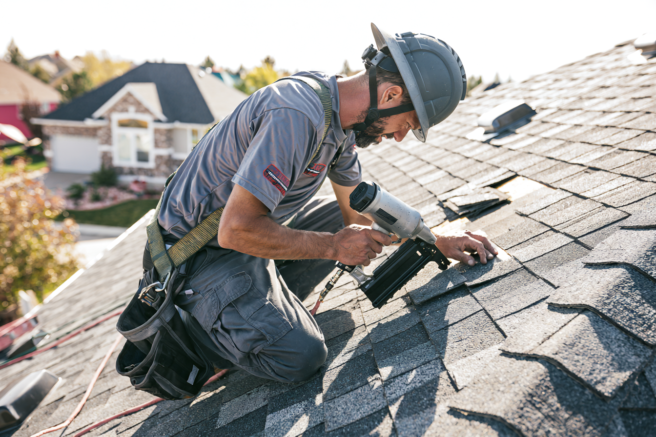 A man wearing a safety helmet and work clothes is installing or repairing shingles on a residential roof using a pneumatic nail gun.
