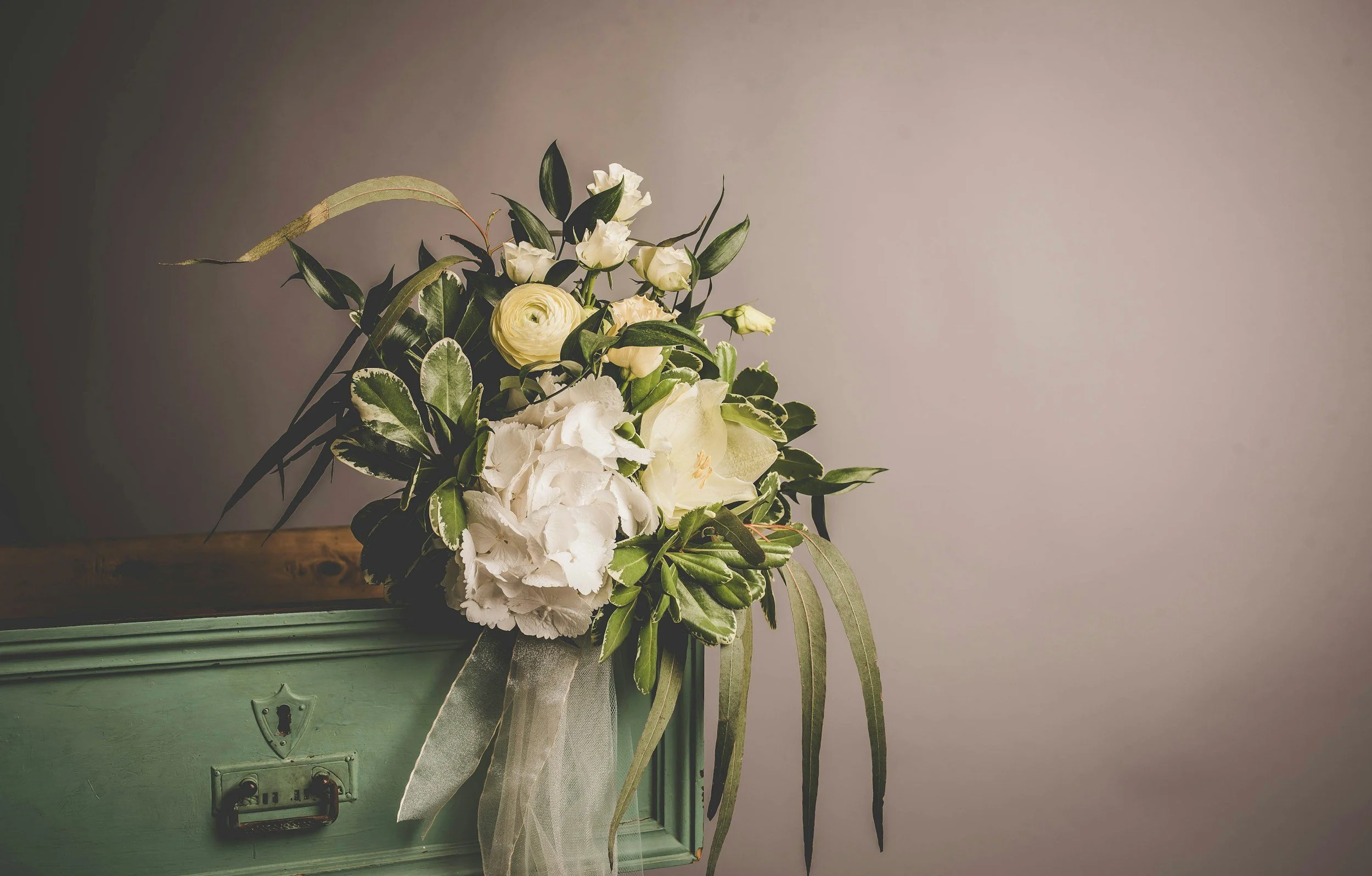 A floral arrangement with white flowers and green leaves, attached to a vintage light green wall-mounted mailbox with a shield-shaped emblem.