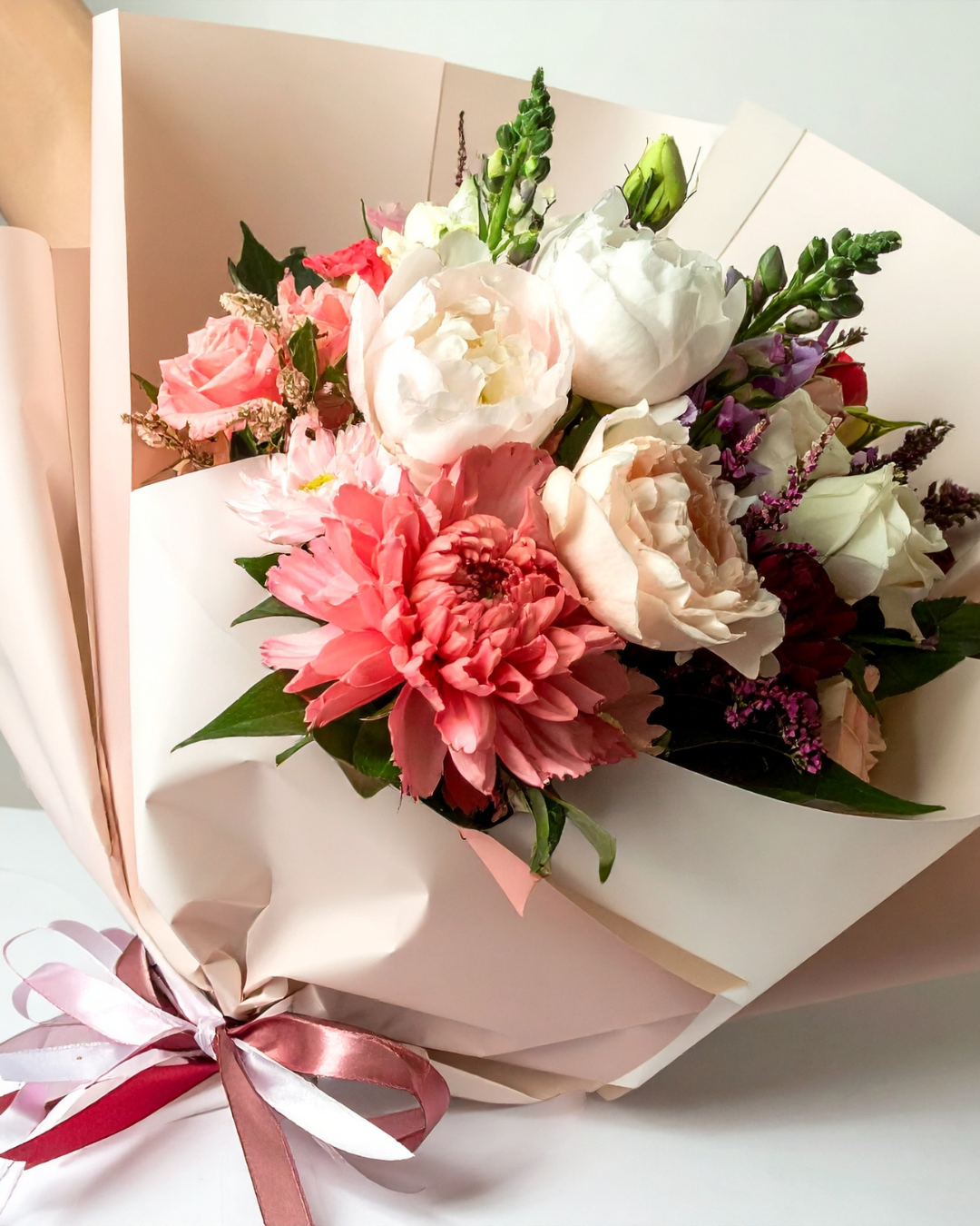 A bouquet of mixed flowers including pink, white, and purple blooms, wrapped in cream paper with a pink ribbon tied around it.