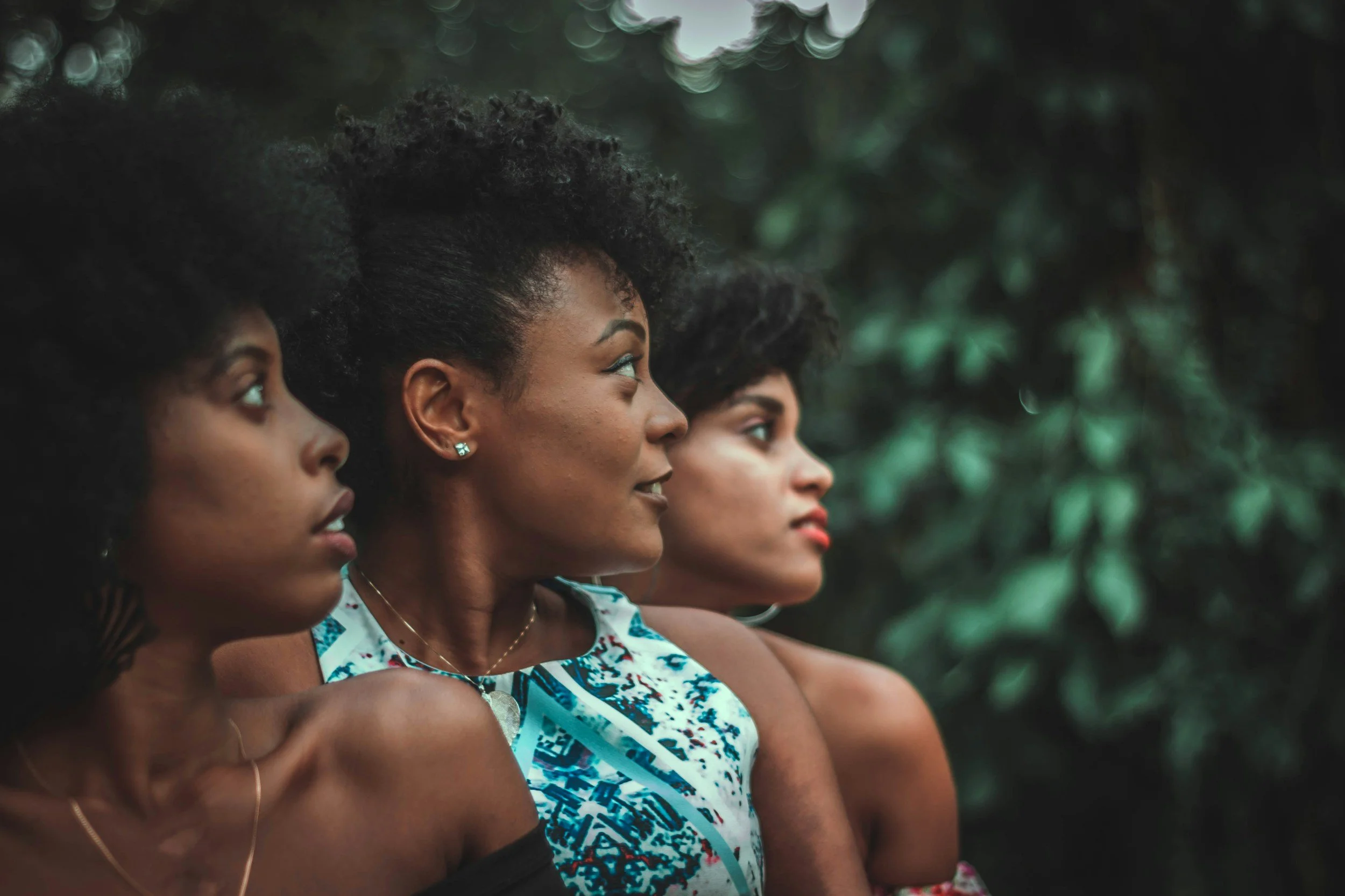 Profile view of four women with dark curly hair, wearing sleeveless dresses, standing outdoors in front of blurred greenery.