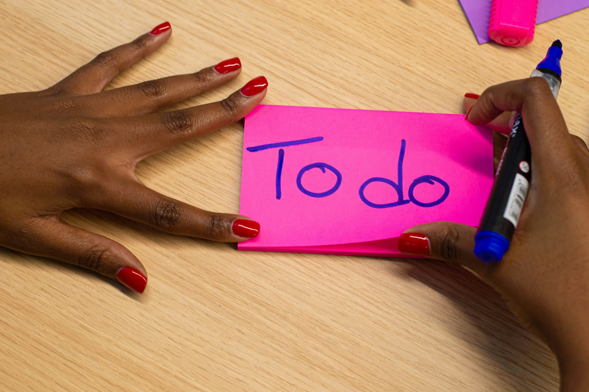 A person with red painted nails signs a pink note with a blue marker that reads '✏️ do' at a wooden table, with a pink highlighter, purple paper, and a black marker nearby.