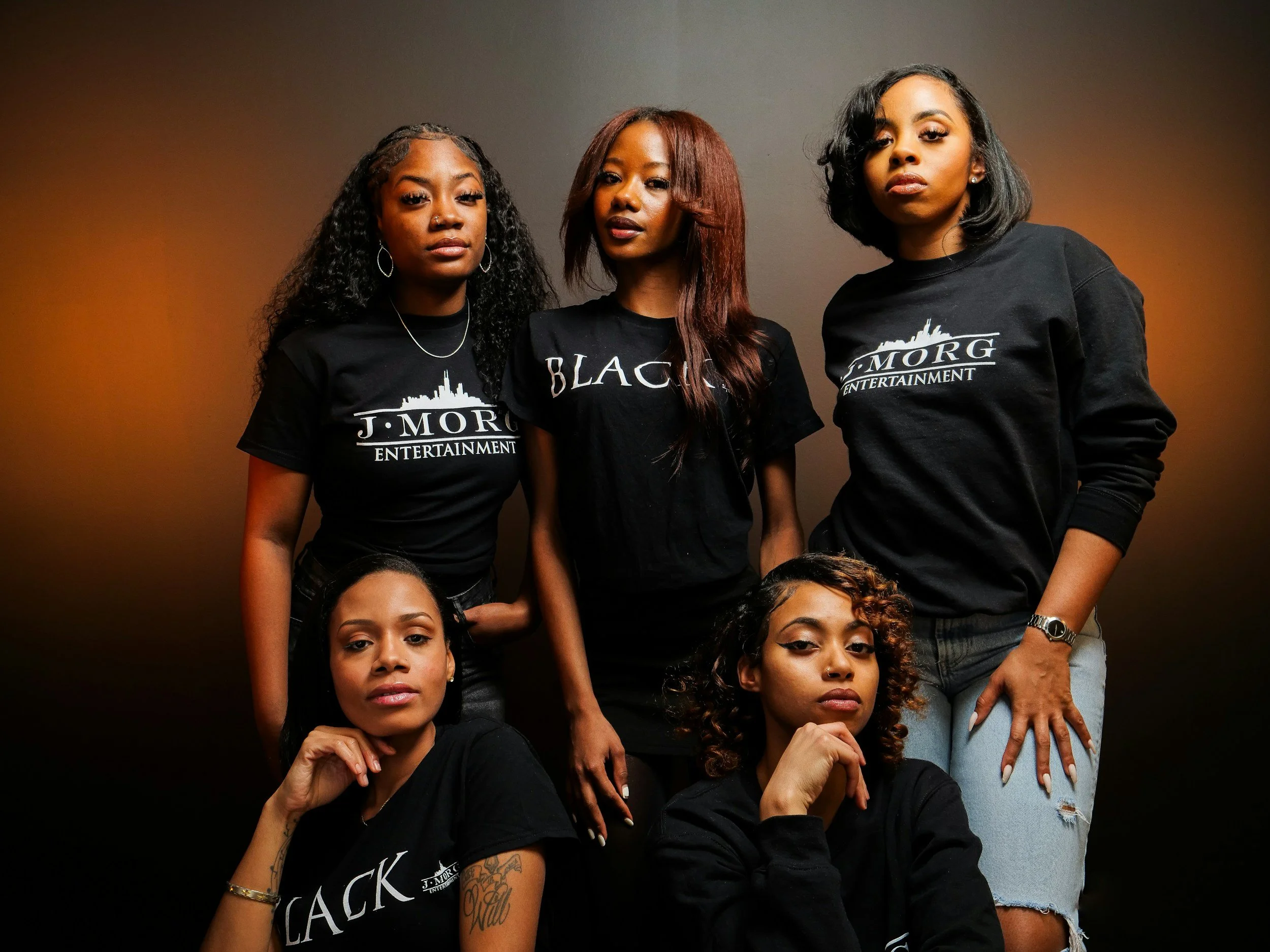 Therapy Groups for women posing for a photo against a plain wall, wearing black T-shirts with 'J. MORG ENTERTAINMENT' printed on them.