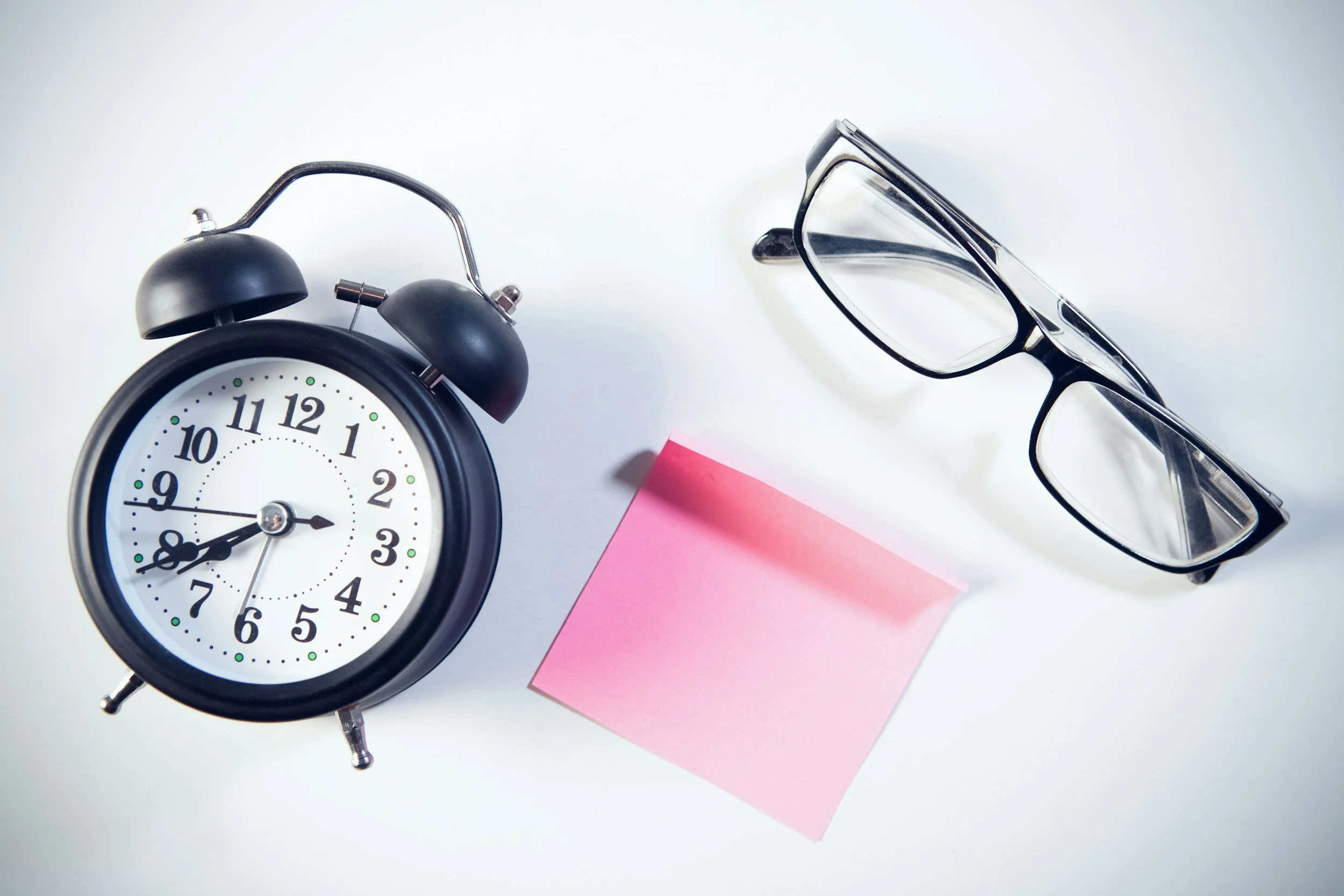 Black vintage alarm clock showing 8:45, black-rimmed eyeglasses, a pink sticky note, and a piece of paper on a white surface.