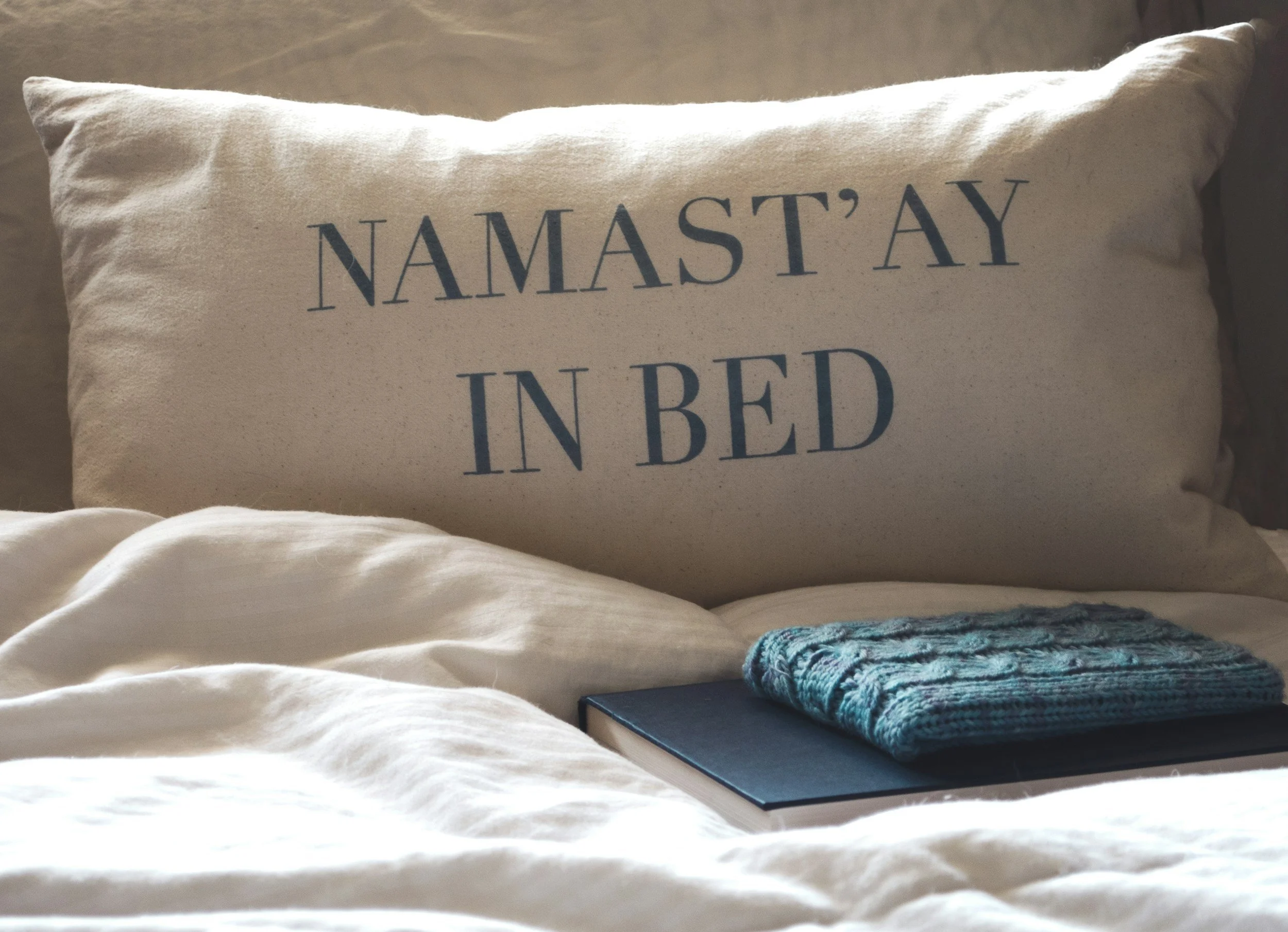 A decorative pillow with the phrase 'Namast'ay in bed' printed on it, placed on a bed with a blanket and a folded knit blanket nearby.
