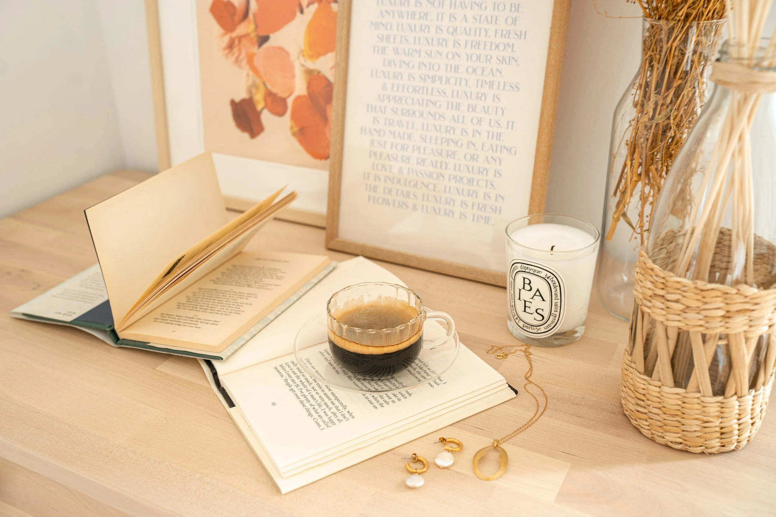 A cozy workspace with open books, a cup of coffee, and various decorative items. There's a framed quote about luxury, a candle, a jewelry piece, and a woven basket on a light wooden table.  Tools to combat signs of burnout in women.
