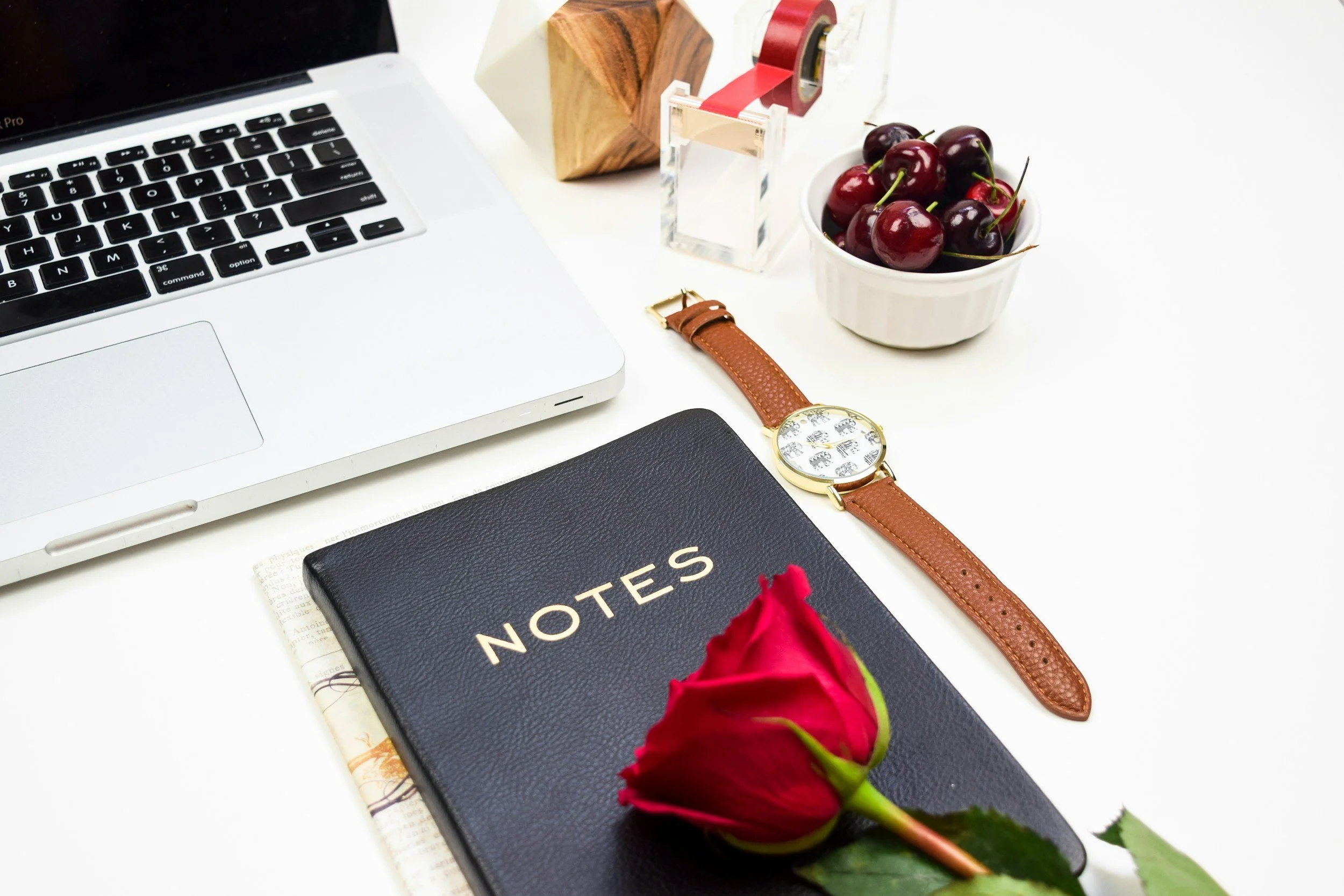 A cluttered white desk with a silver Apple MacBook, a rose, a notebook titled "NOTES," a black textured planner, a rolling pin, a wooden geometric object, a tape dispenser with transparent tape, ready to answer Therapy Questions
