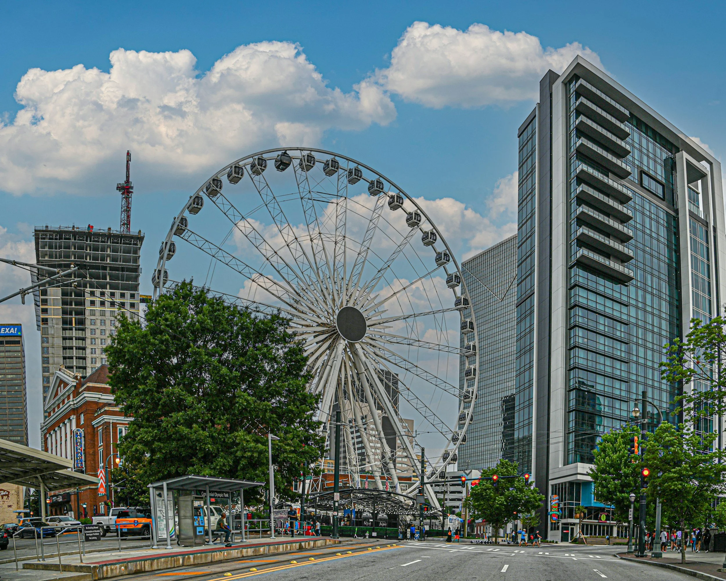 A GA therapy cityscape featuring a large Ferris wheel, modern glass high-rise buildings, and a partially constructed building in the background under a partly cloudy sky.