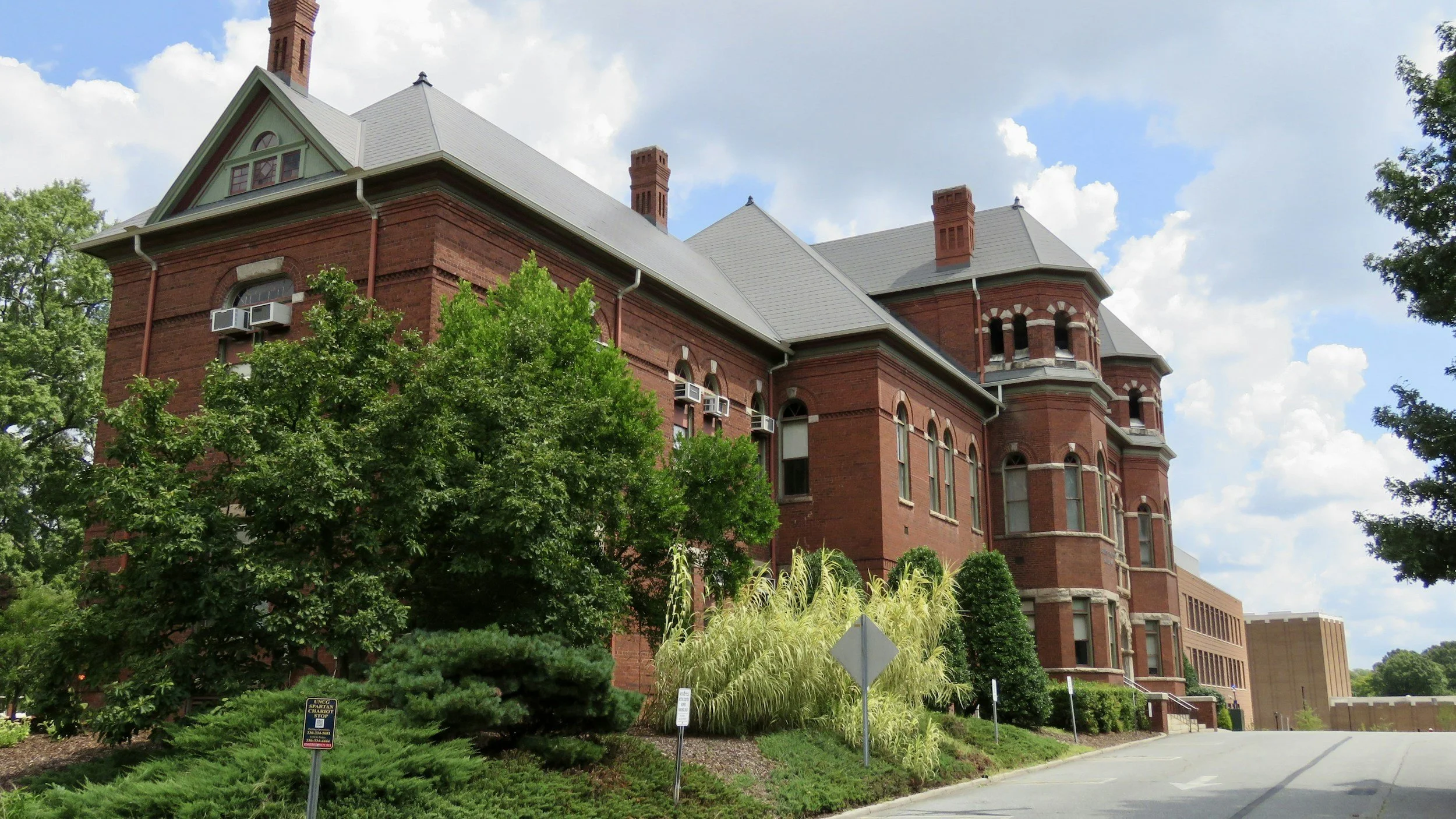 A large, historic red brick building with a cloudy blue sky above and lush green trees in front.