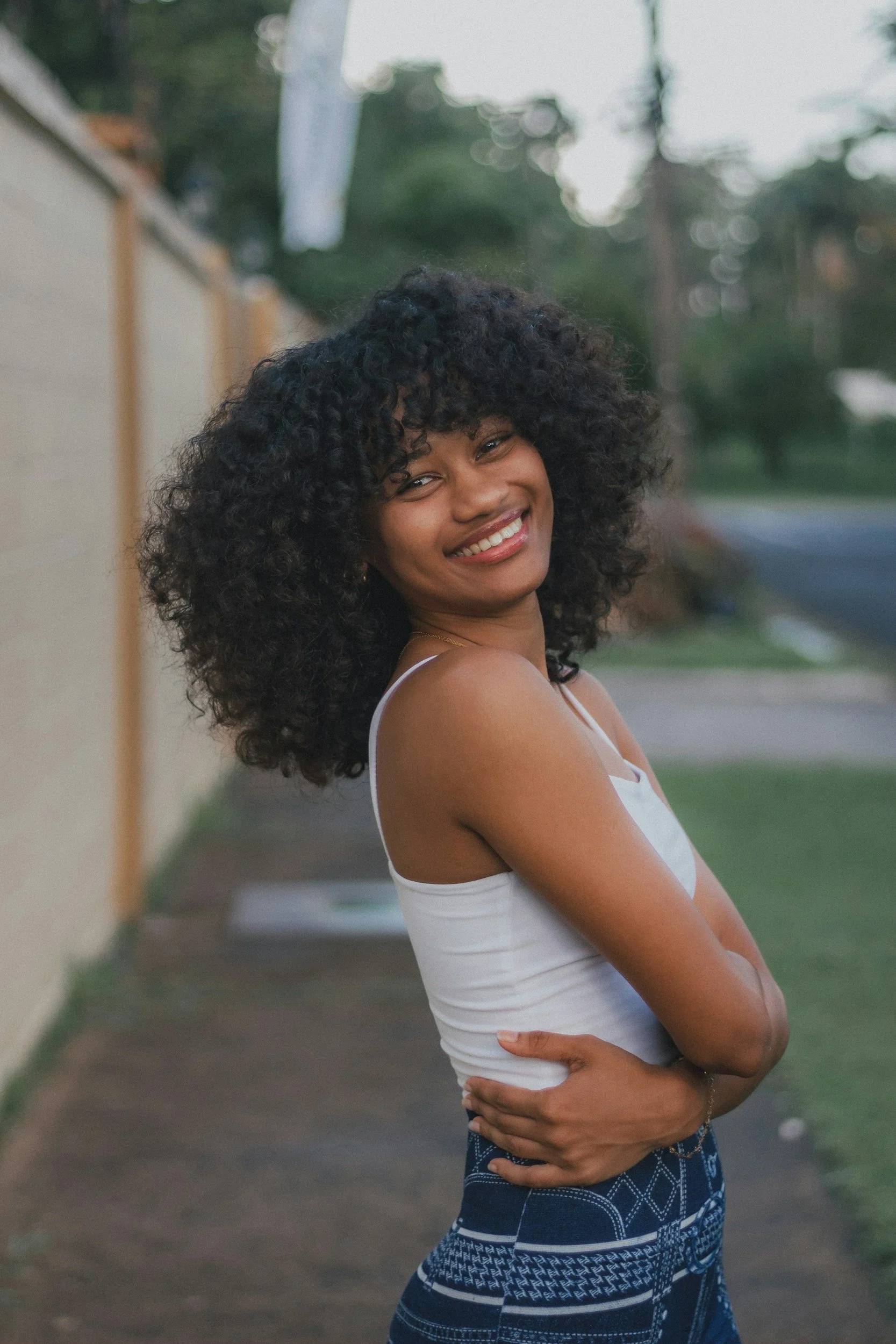 A woman with curly black hair smiling outside, wearing a white tank top and patterned blue pants, standing near a wall with trees in the background.