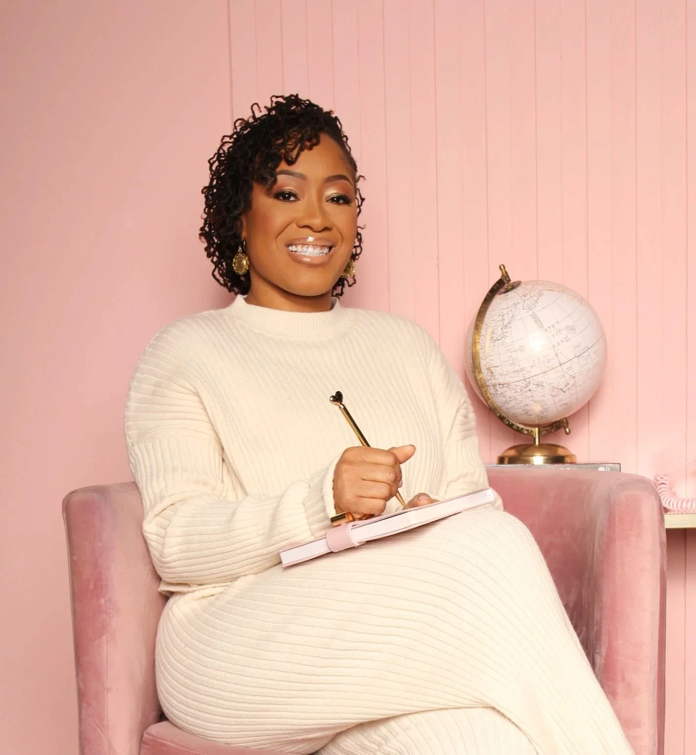 A woman sitting in a pink velvet chair, smiling and holding a notebook and pen, with a globe on a table behind her, all against a pink wall.