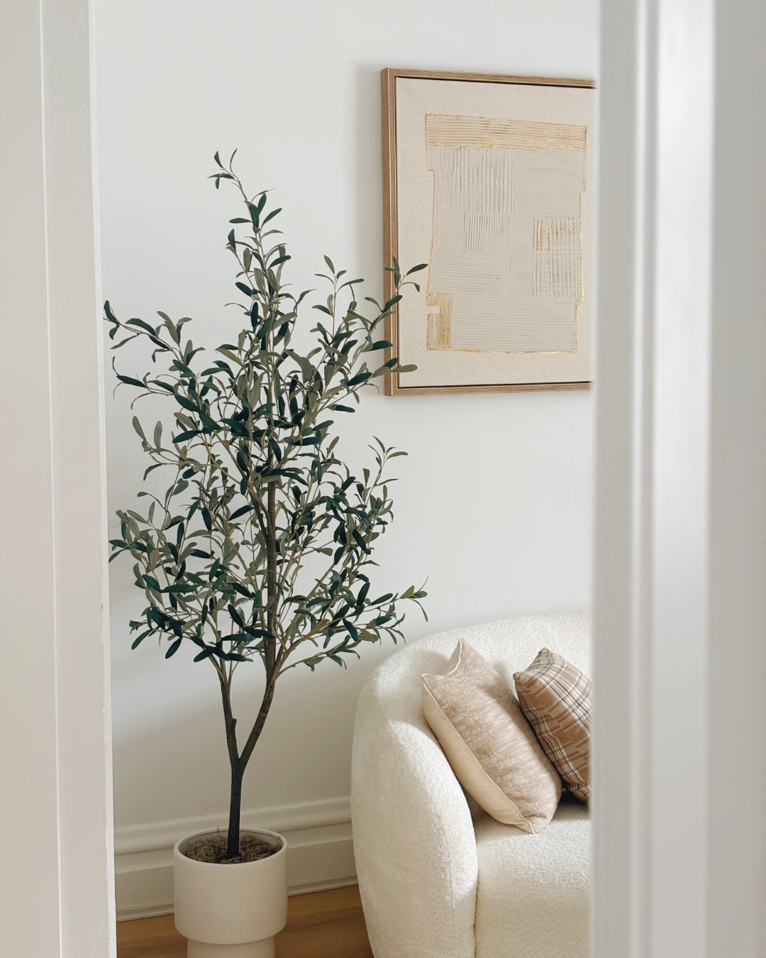 A cozy corner with a tall potted plant, a white textured armchair with two throw pillows, and a framed abstract art piece on the wall.