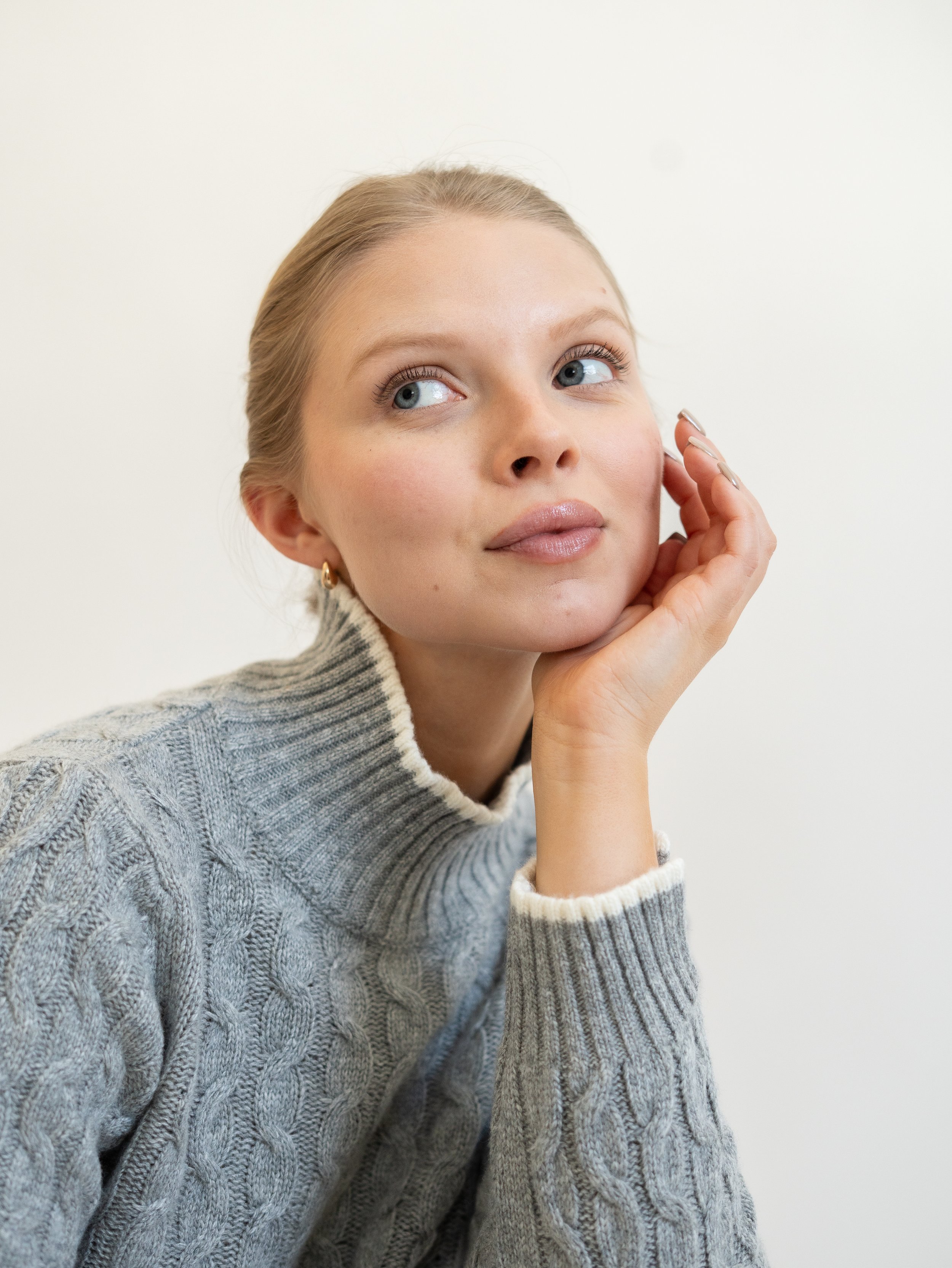 A woman with blonde hair and blue eyes wearing a gray cable-knit sweater, resting her chin on her hand against a plain white background.
