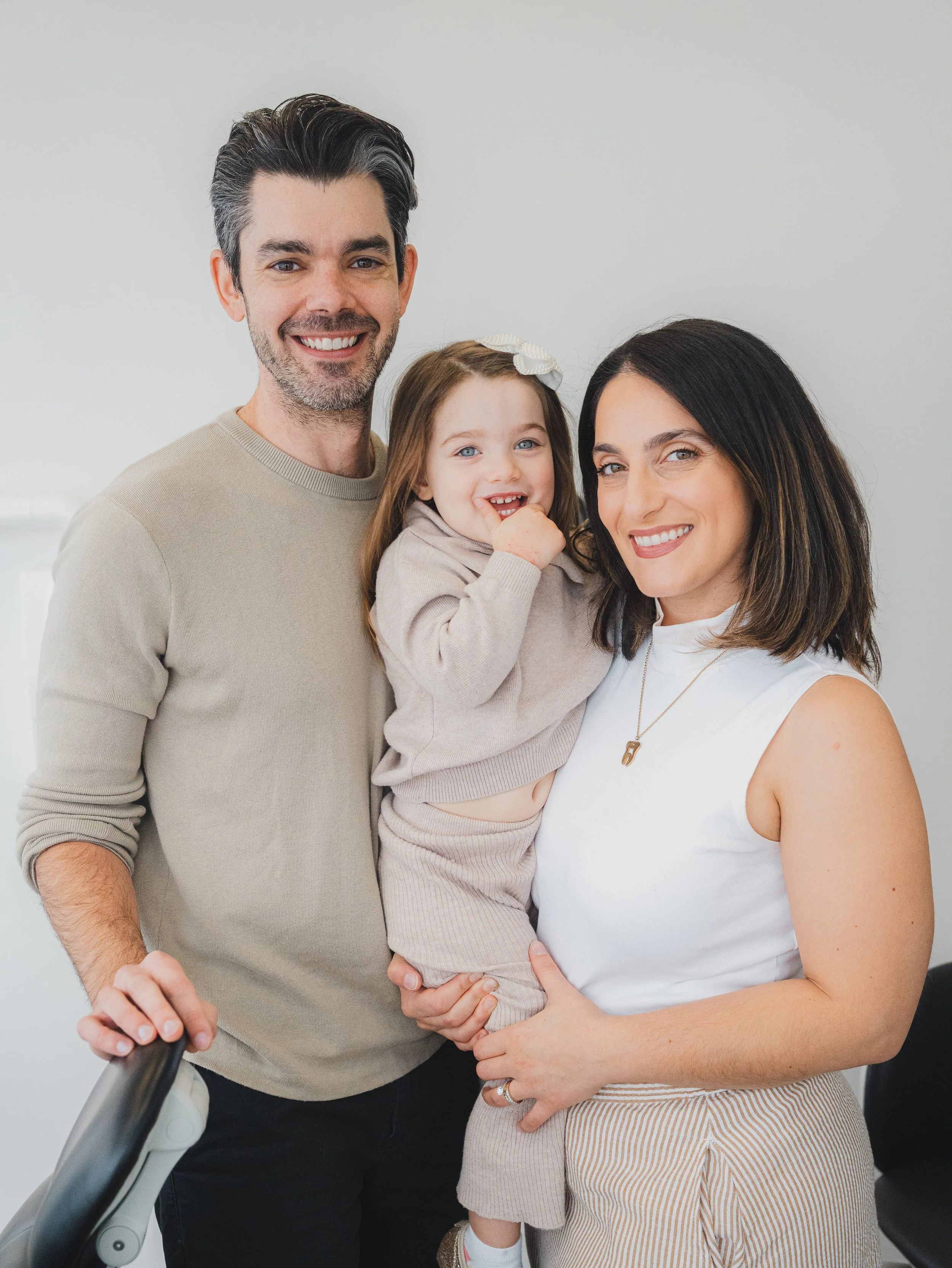 A happy family of three: a man, a woman, and a young girl, standing together indoors in front of a plain white wall, smiling at the camera.