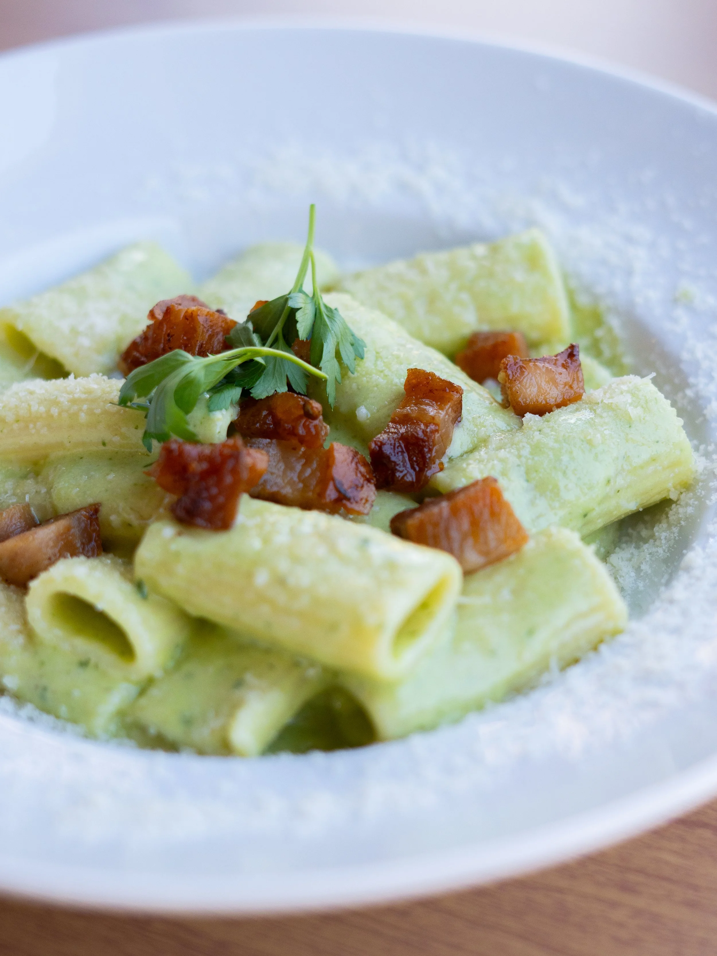 Close-up of fresh pasta ribbons topped with cooked bacon pieces and garnished with a sprig of parsley, served in a white bowl.