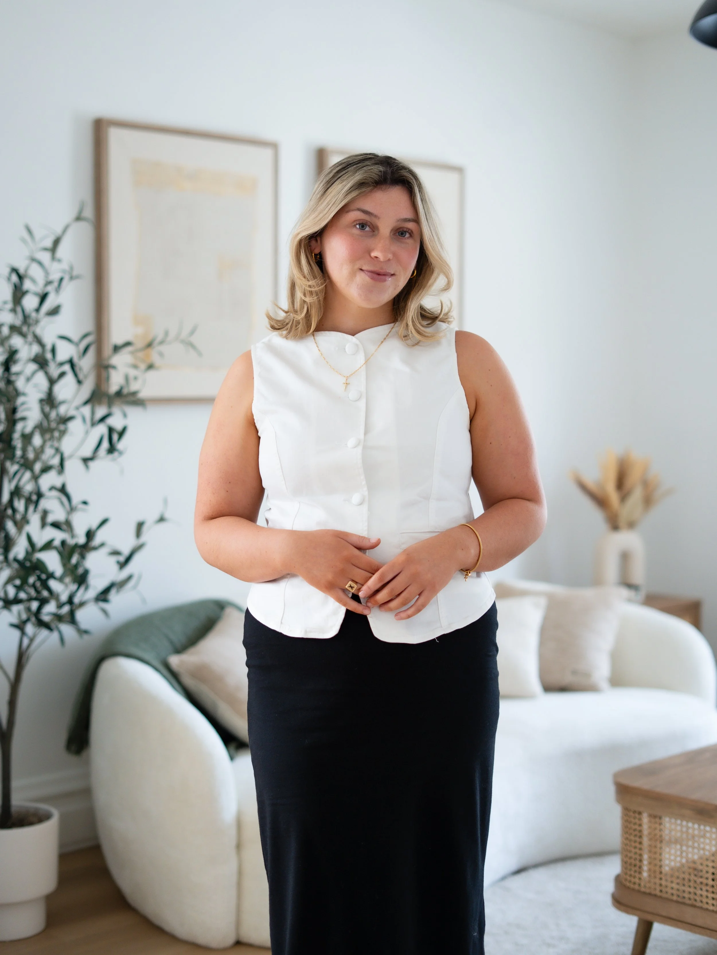 A woman with blonde hair wearing a white sleeveless top and a long black skirt, standing in a living room with a white couch, a potted plant, and framed artwork on the wall.