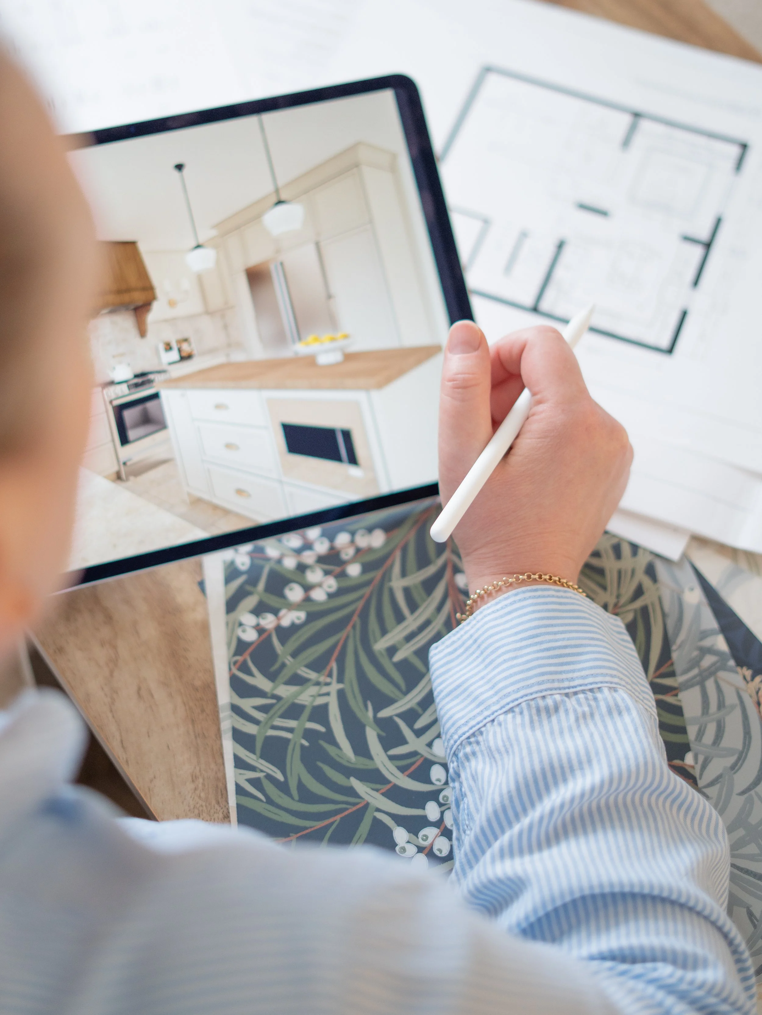 Person in blue and white striped shirt using a stylus on a tablet in a bright, modern kitchen with white cabinets and pendant lights.