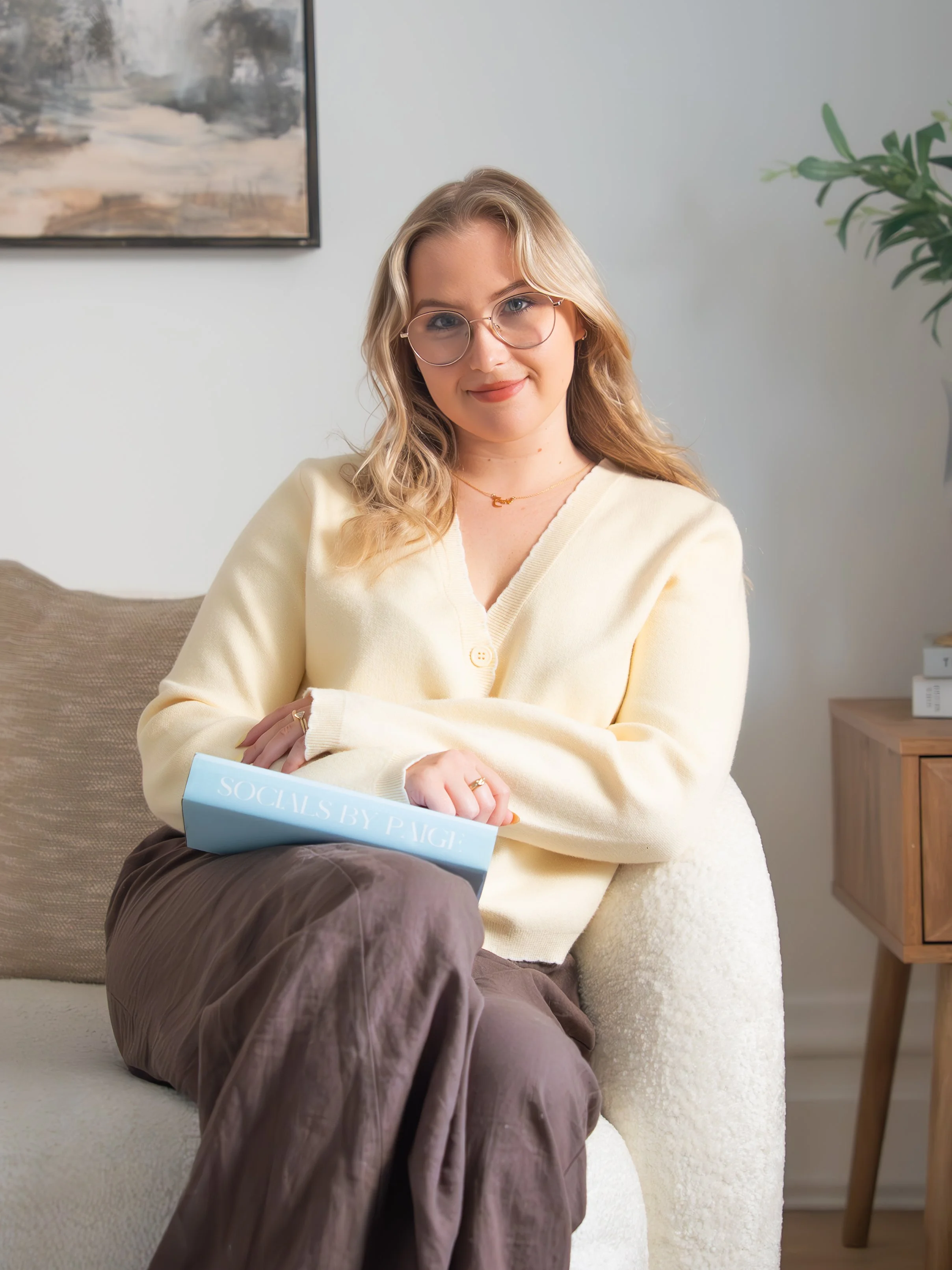 Young woman sitting on a beige couch holding a book titled 'Socials by P. PAGE' with a neutral expression, wearing glasses, a cream sweater, and brown pants, in a room with a painting on the wall and a green plant.