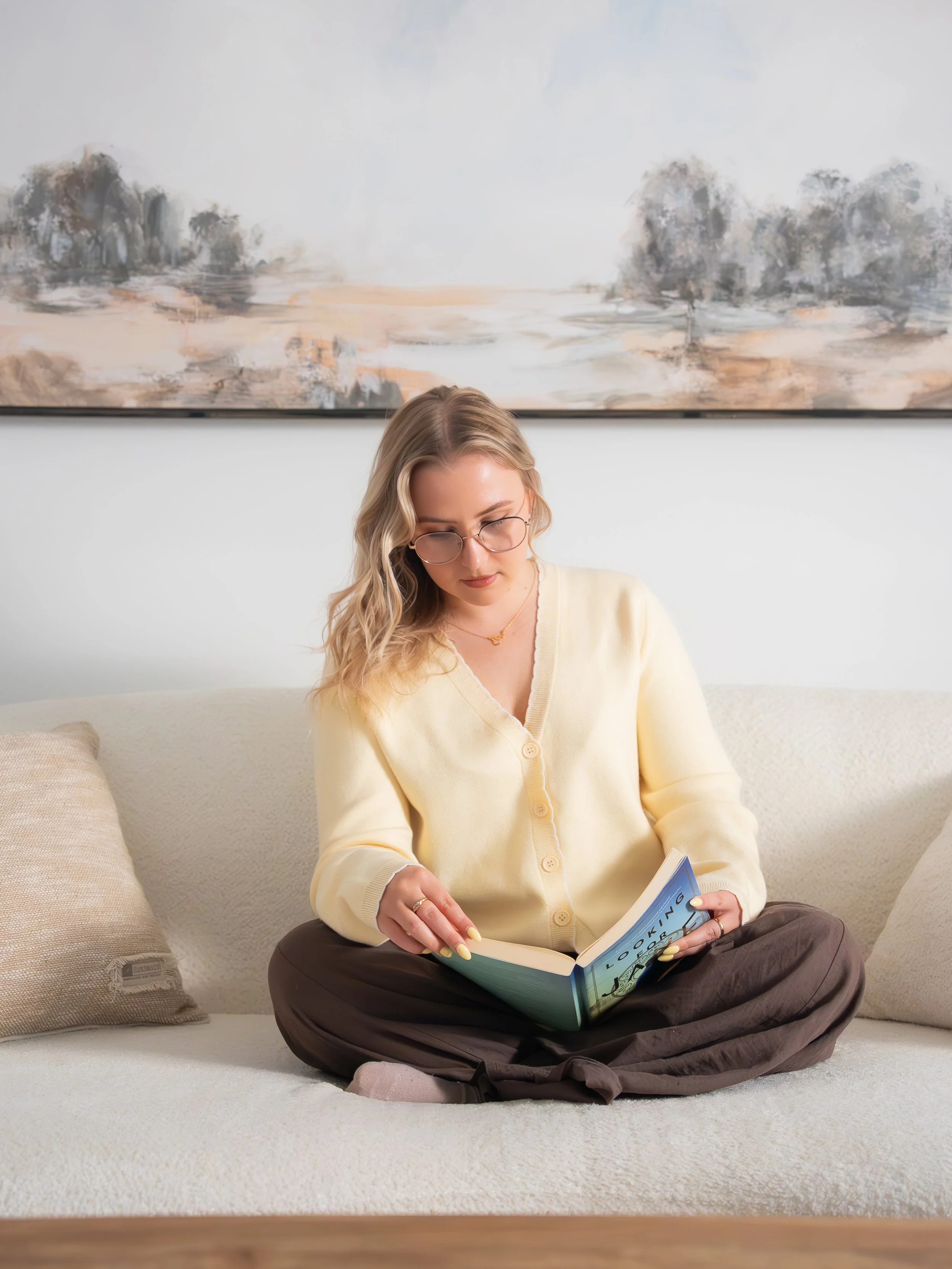 A young woman with blonde wavy hair and glasses sitting on a white couch, reading a book with a blue cover, dressed in a pale yellow cardigan and brown pants, in a modern living room with a large abstract painting hanging on the white wall behind her.