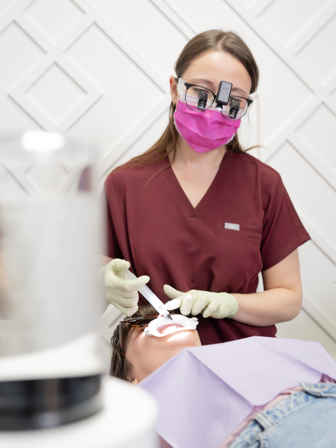 A dentist wearing pink mask and magnifying glasses with dental tools working on a patient's open mouth in a dental office.
