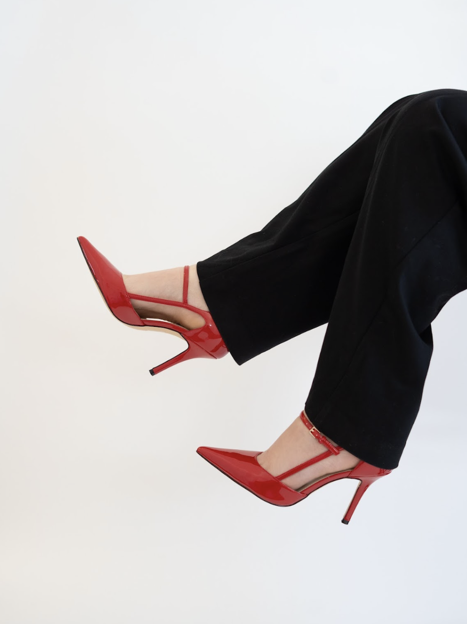 Close-up of a person's legs wearing black pants and red high-heeled shoes, with legs crossed at the ankles, against a plain white background.