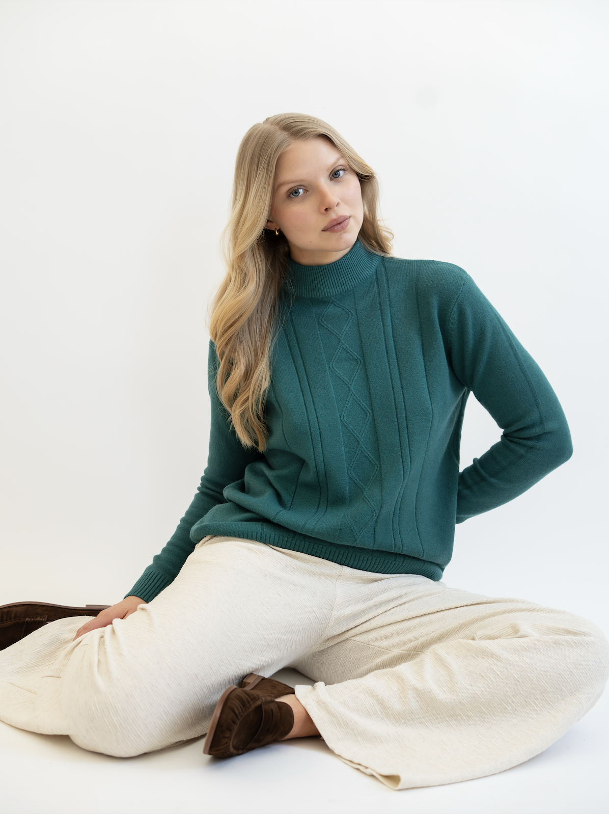 A woman with blonde hair, blue eyes, and fair skin, sitting cross-legged in front of a plain white background, wearing a teal sweater and cream-colored pants.