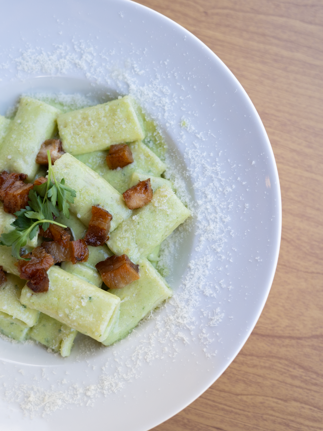 Plate of green tortellini pasta garnished with cooked bacon bits and a sprig of parsley, with grated cheese around the edge of the white plate, on a wooden surface.