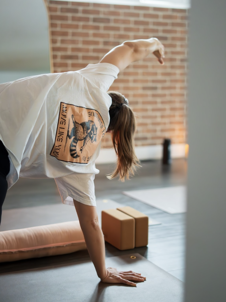 A woman doing yoga in a studio, in a side bend pose with her right arm reaching over her head, wearing a white t-shirt with a graphic of a tiger and the words 'Loves You' on the back.