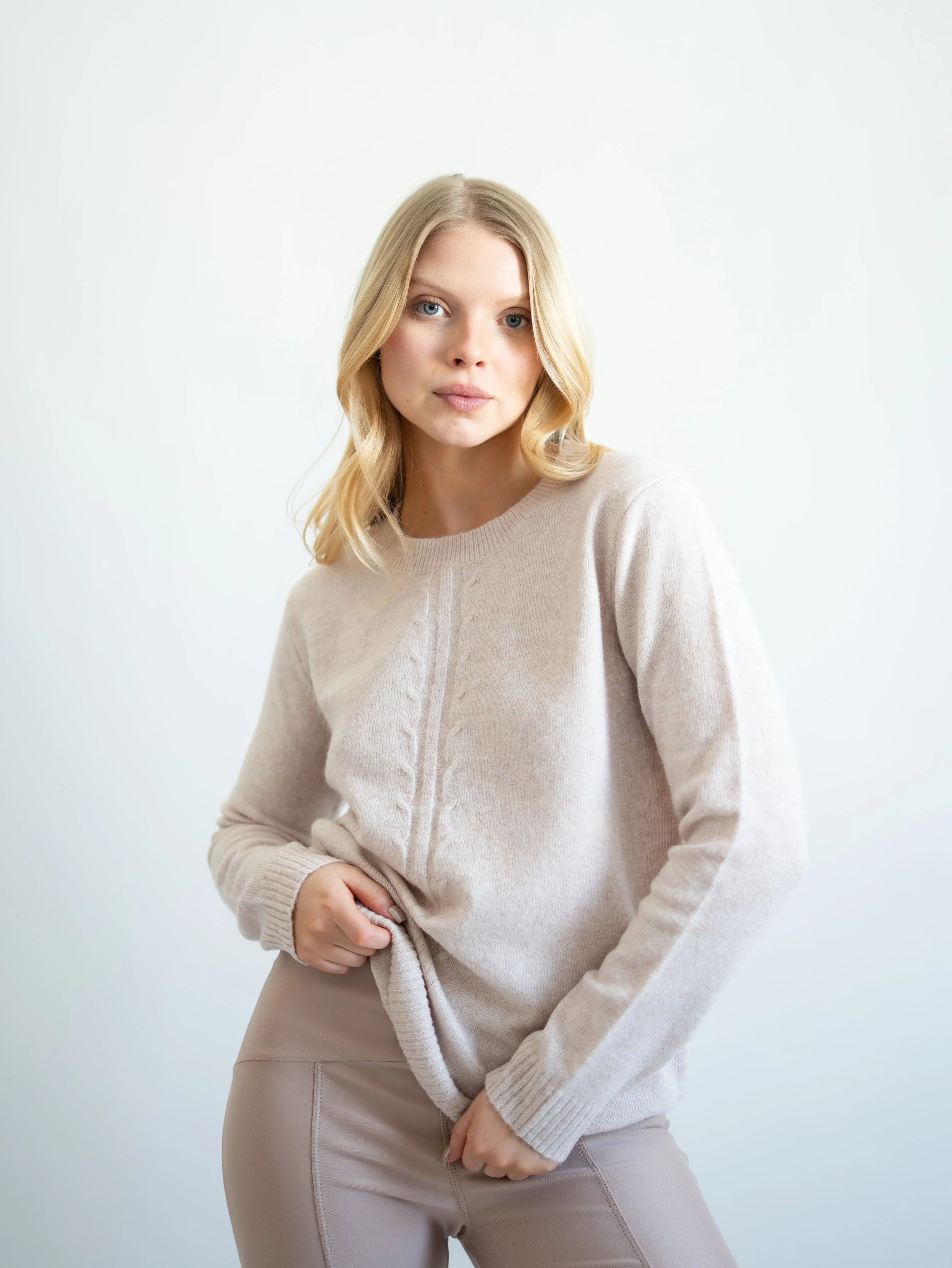 A woman with blonde hair wearing a beige sweater and beige pants standing against a plain white background.