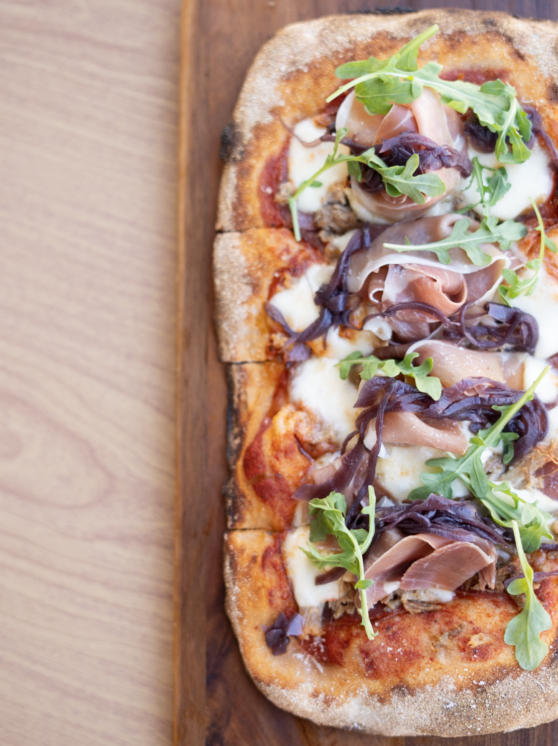 A rectangular pizza topped with prosciutto, arugula, black olives, and melted cheese on a wooden serving board.