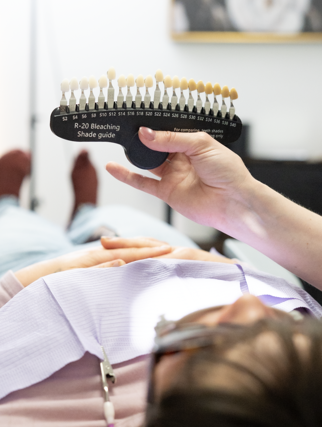 Person holding a shade guide for teeth with a patient lying down in a dental chair in the background.