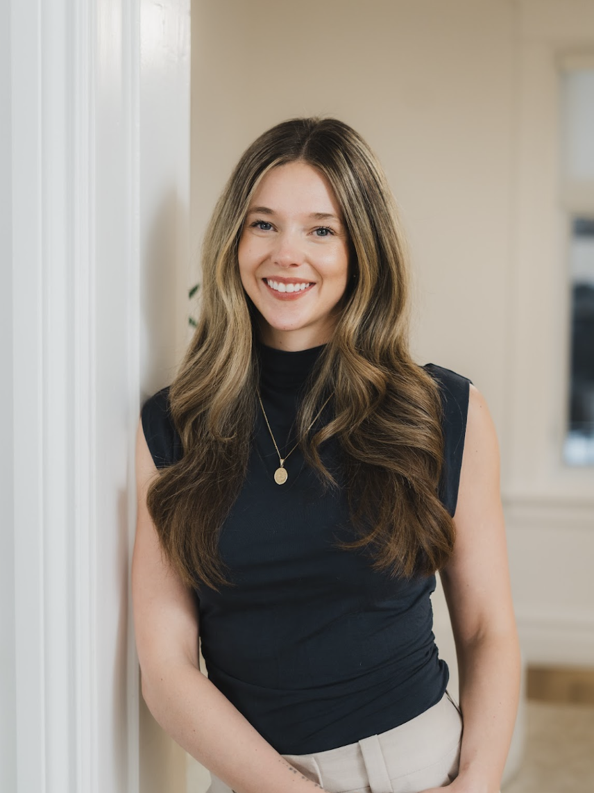 A young woman with long wavy brown hair and a bright smile, wearing a black sleeveless top and a necklace, standing indoors next to a white wall.