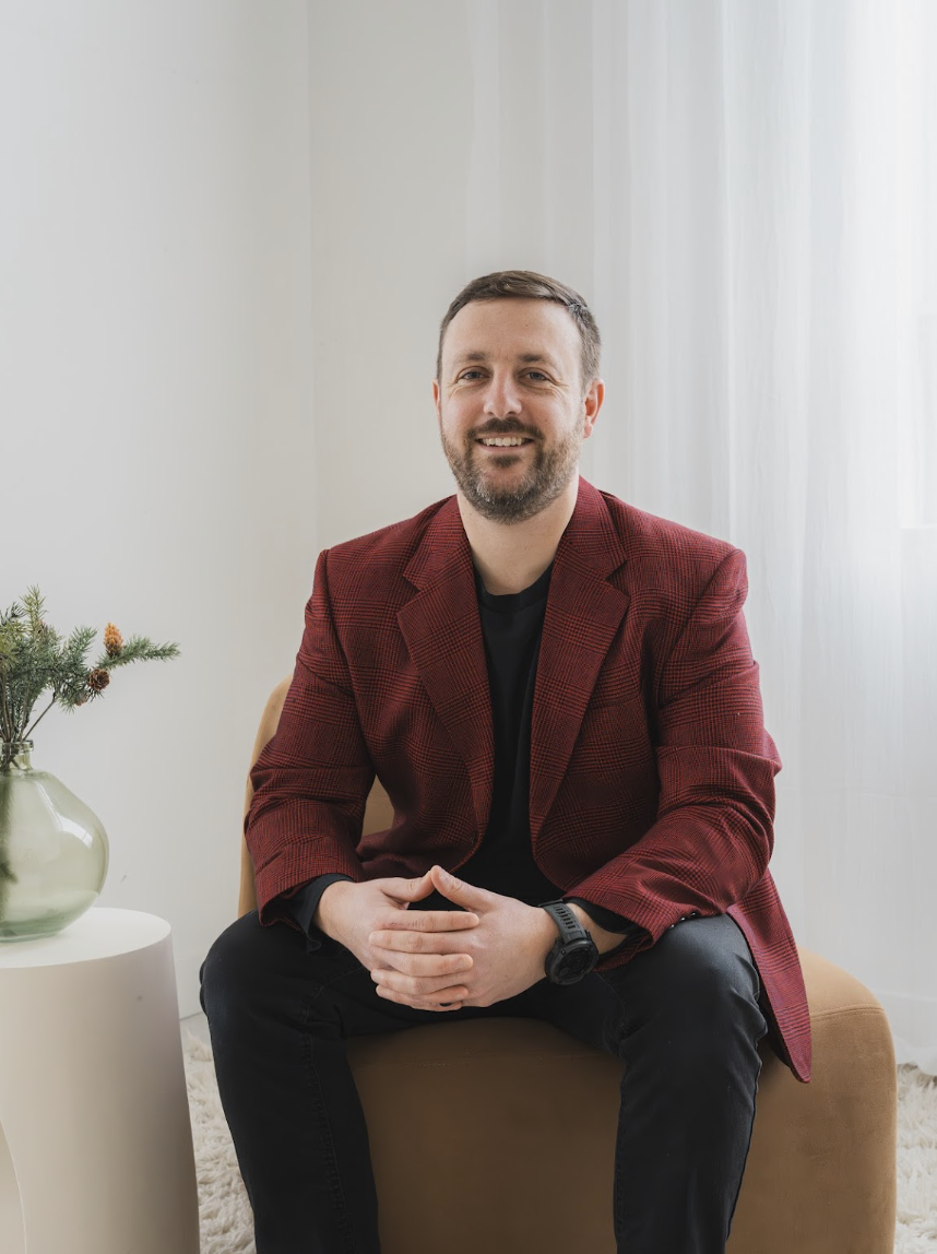 A man with a beard and short hair, wearing a red blazer and black shirt, sitting on a tan chair in a room with white curtains and a white wall, smiling at the camera. A white side table with a green vase and pine branch is next to him.