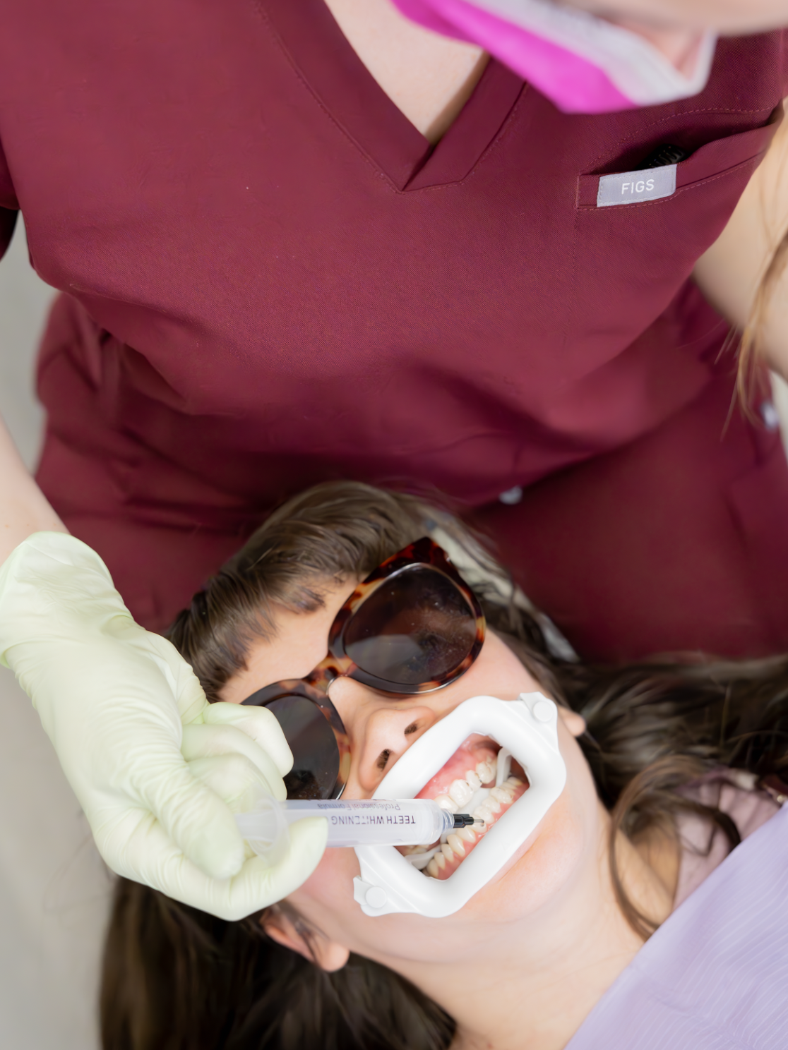 A woman at the dentist's office receiving dental treatment while lying back, wearing sunglasses and a dental shade. A dental professional is using a syringe marked 'Teeth Whitening' near her mouth.