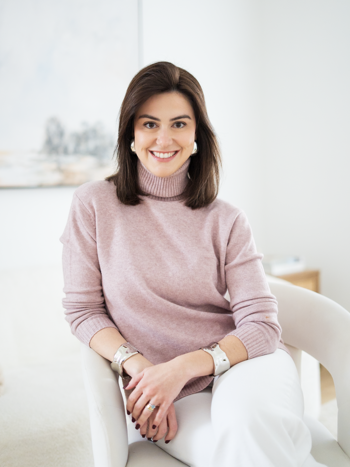 A woman smiling while sitting in a white armchair, wearing a light pink turtleneck sweater, white pants, and silver jewelry, in a light-colored room.