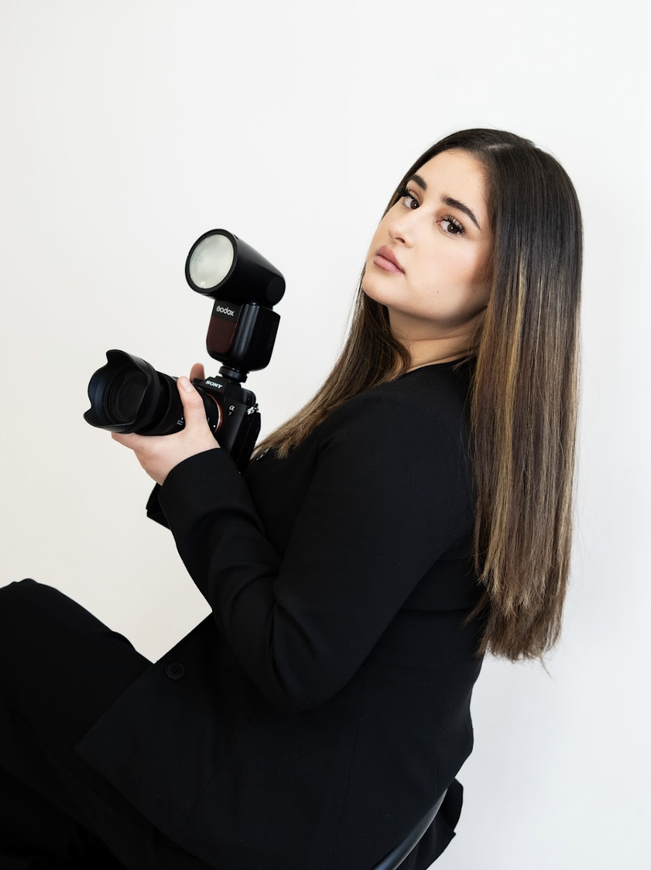 A young woman with long brown hair, wearing a black blazer, holding a camera with a flash attachment, sitting against a plain white background.