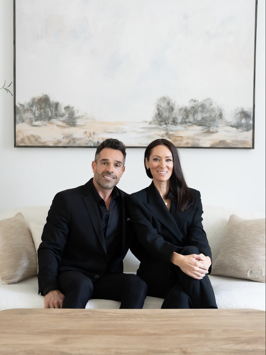 A man and woman sitting on a light-colored sofa in a modern living room with a large abstract landscape painting hanging on the wall behind them. Both are dressed in black suits and smiling.