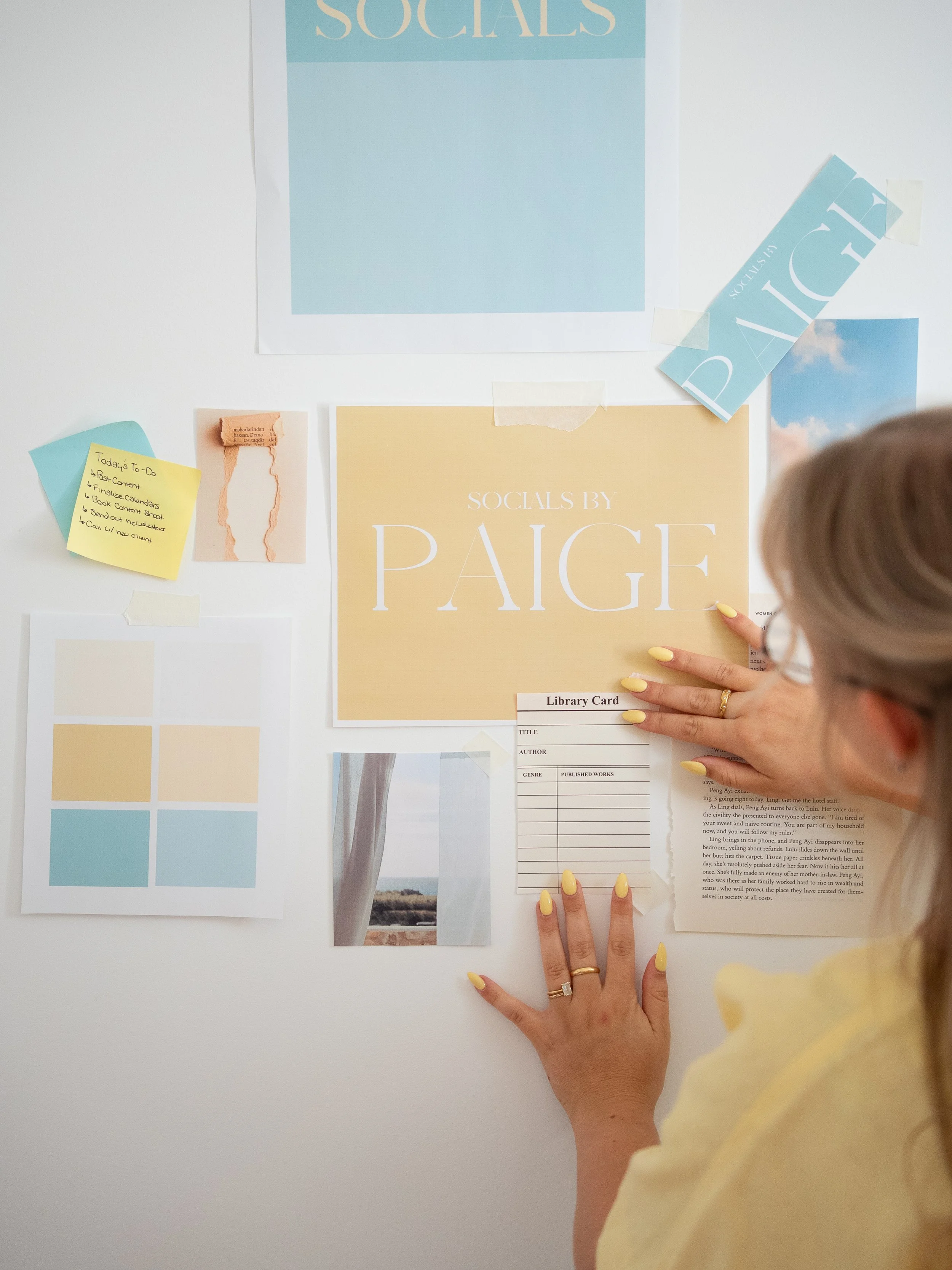 A person with yellow painted nails is placing or pointing at a large beige-colored poster titled 'SOCIALS BY PAGE' on a white wall. The person's hand and part of their face are visible, with their face slightly turned away from the camera.