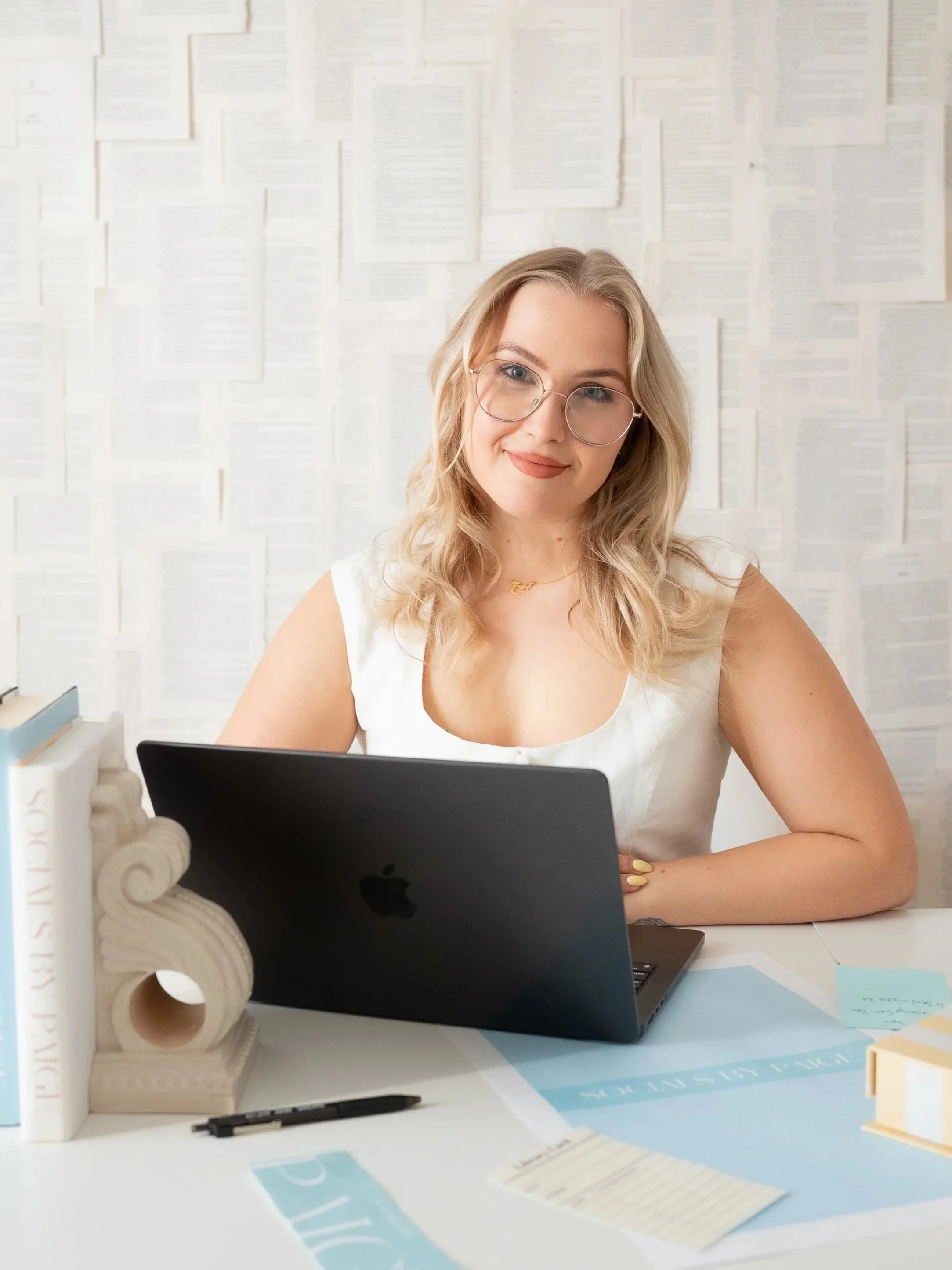 A young woman with blonde hair, glasses, and light makeup, sitting at a desk with a laptop, in front of a wall covered with pages from books or documents and working or studying.