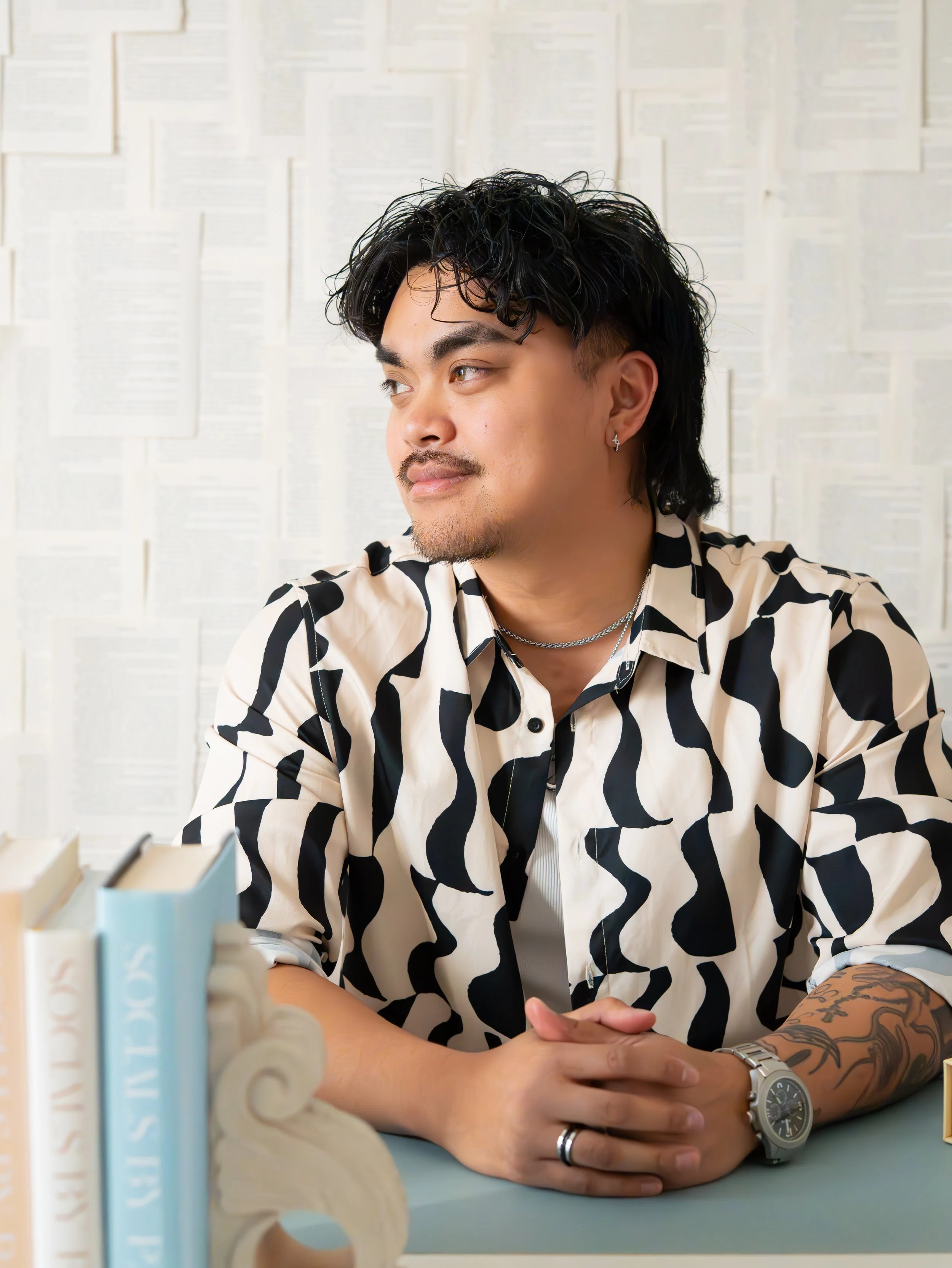 A man with dark curly hair, mustache, and earrings, wearing a patterned shirt, jewelry, and a watch, sitting at a table with his hands folded, in a room with a wall covered in white papers or pages.