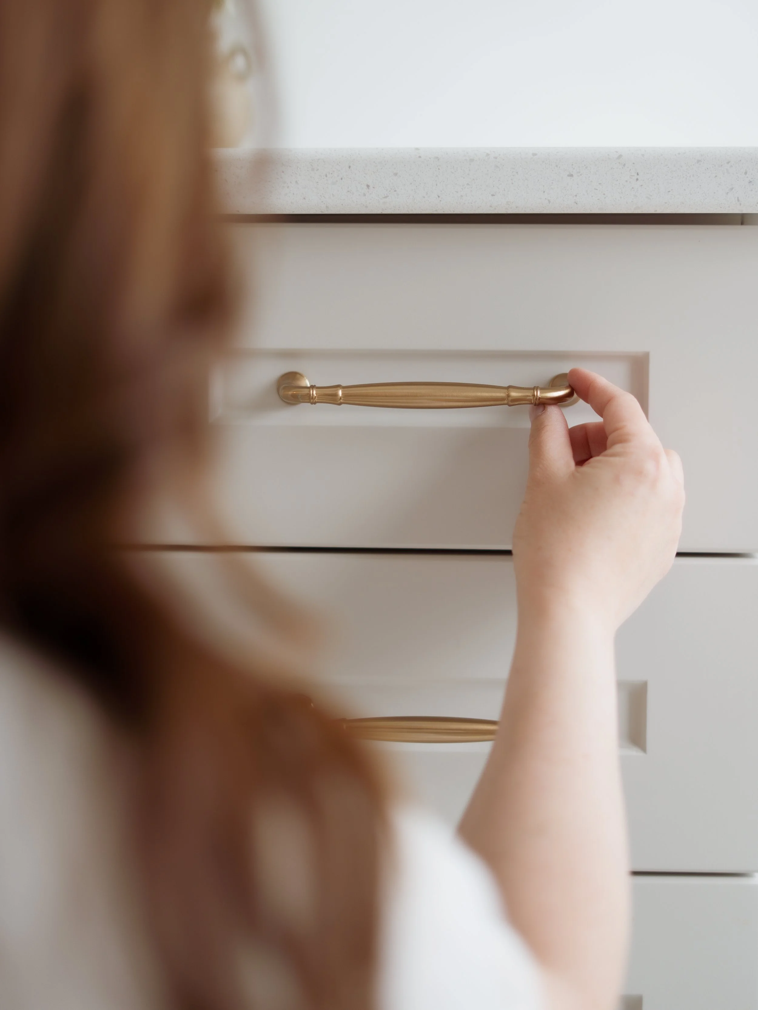 A person with red hair looking at a mirror while holding a gold drawer handle on a white dresser.