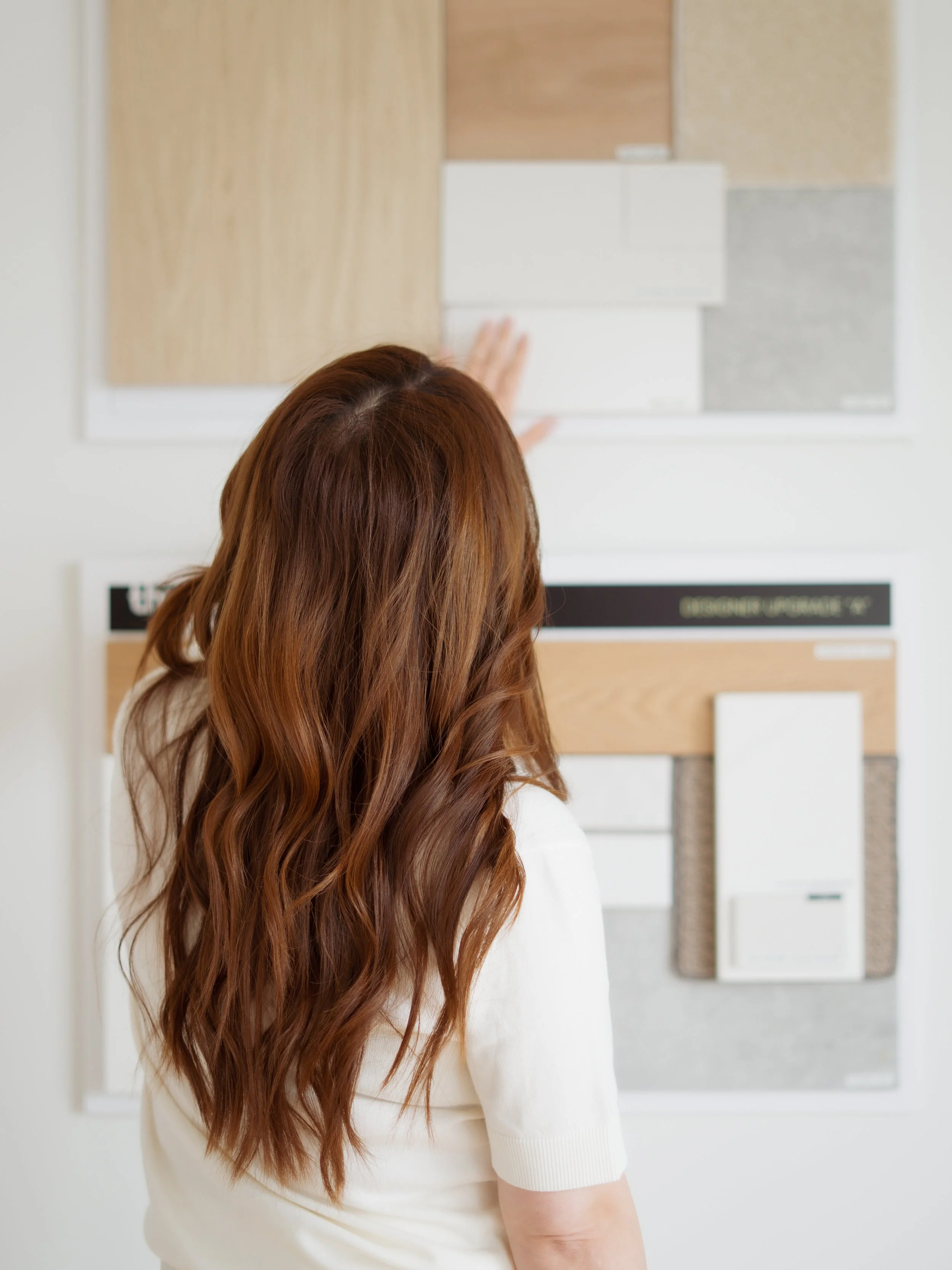 A woman with long, wavy, reddish-brown hair pointing at samples of different wood and material textures displayed on a wall.
