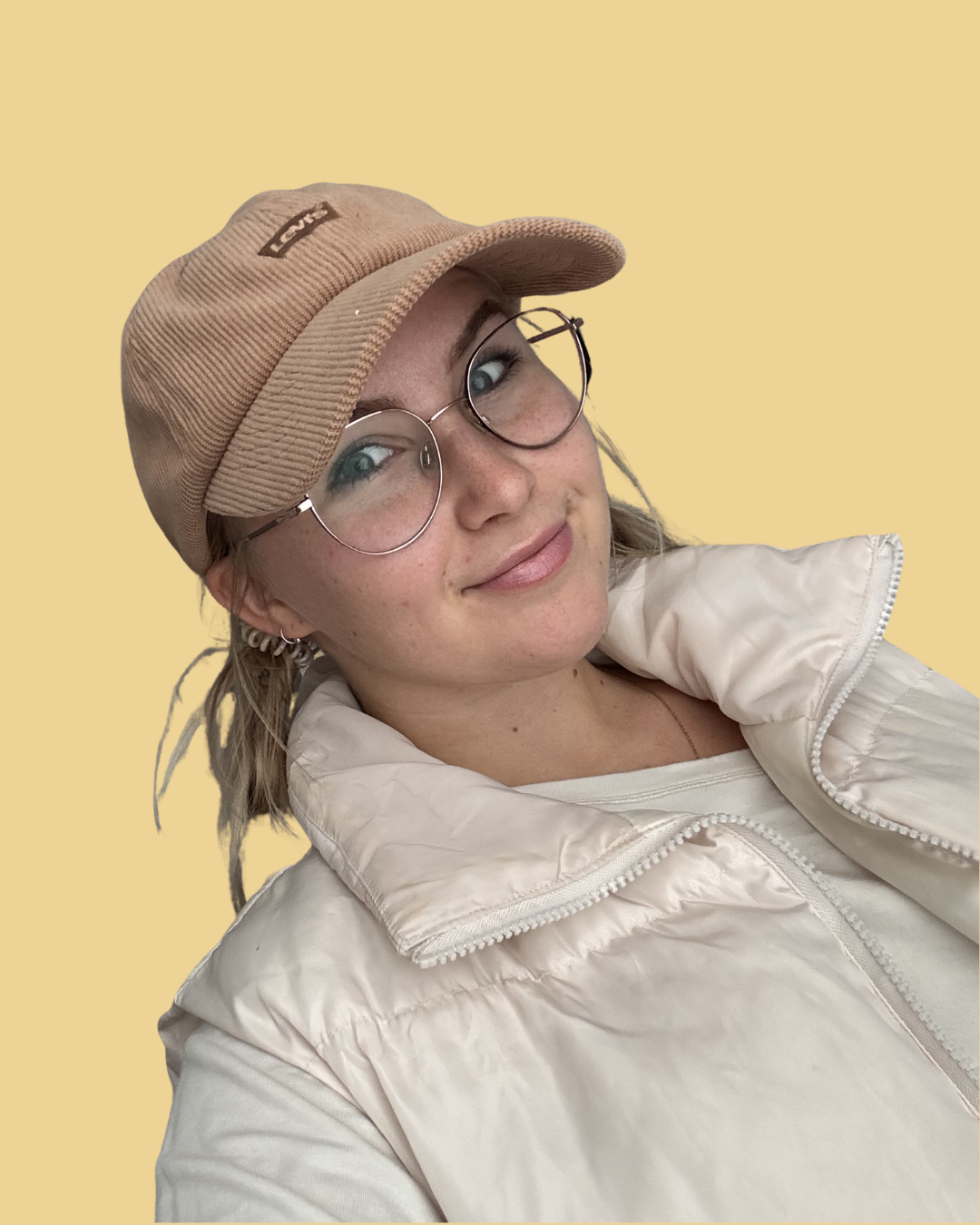 A young woman with glasses, freckles, and a warm smile, wearing a tan cap and a cream-colored puffer jacket, stands against a plain yellow background.