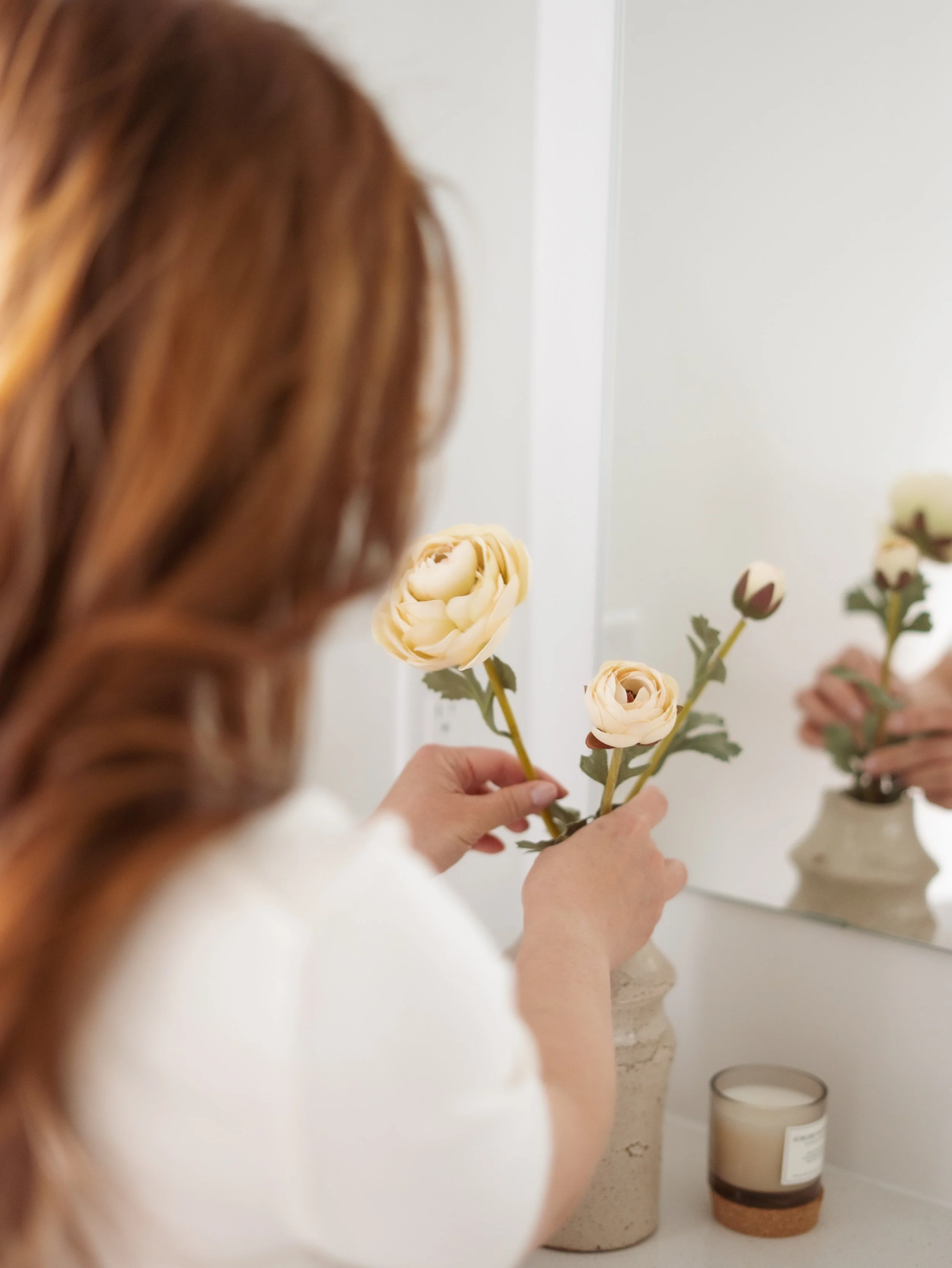 A woman with red hair holding three cream-colored roses in front of a mirror.